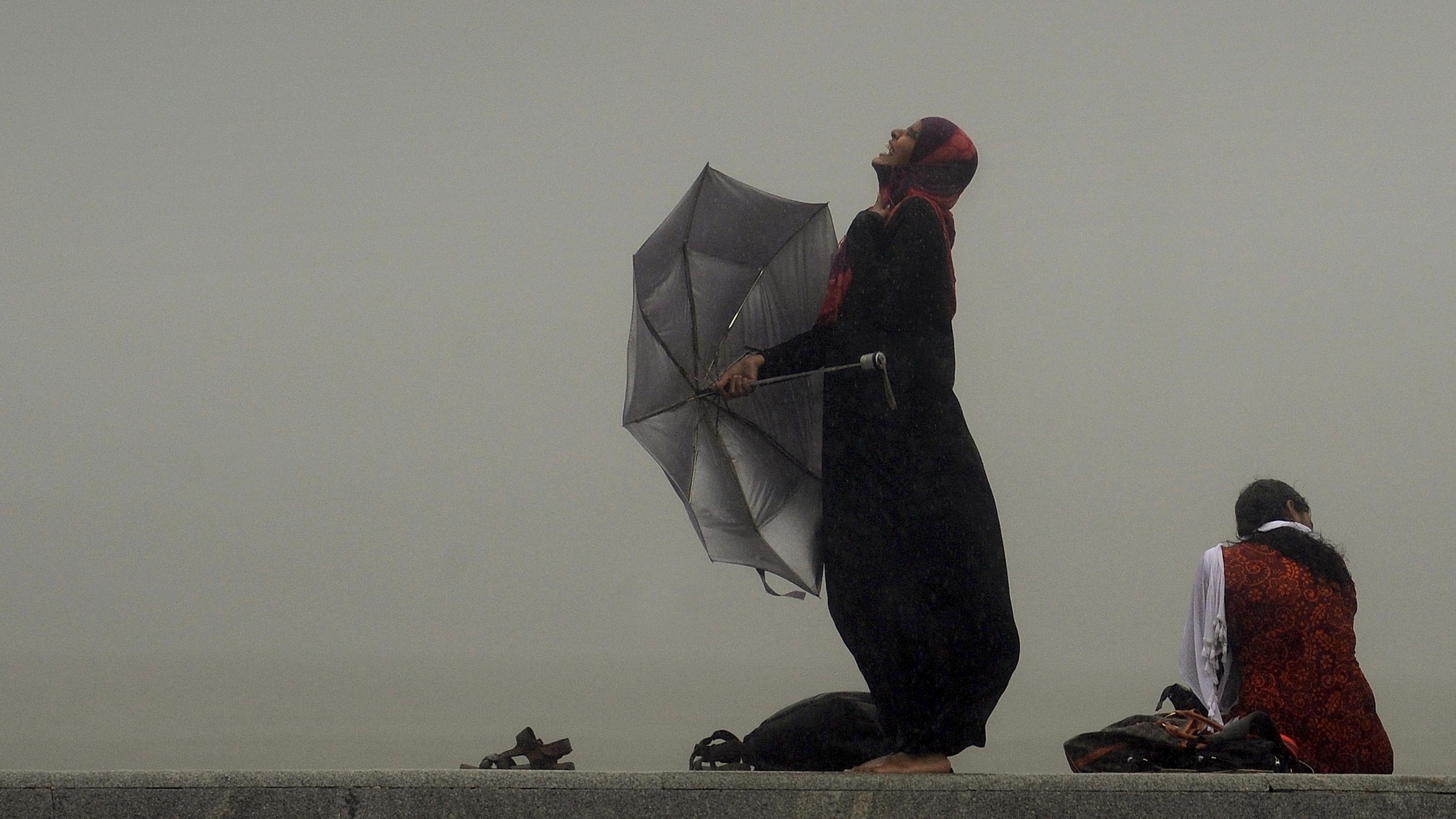 Photo of a woman in the rain holding a broken umbrella, smiling upwards; another sits nearby with a foggy background.