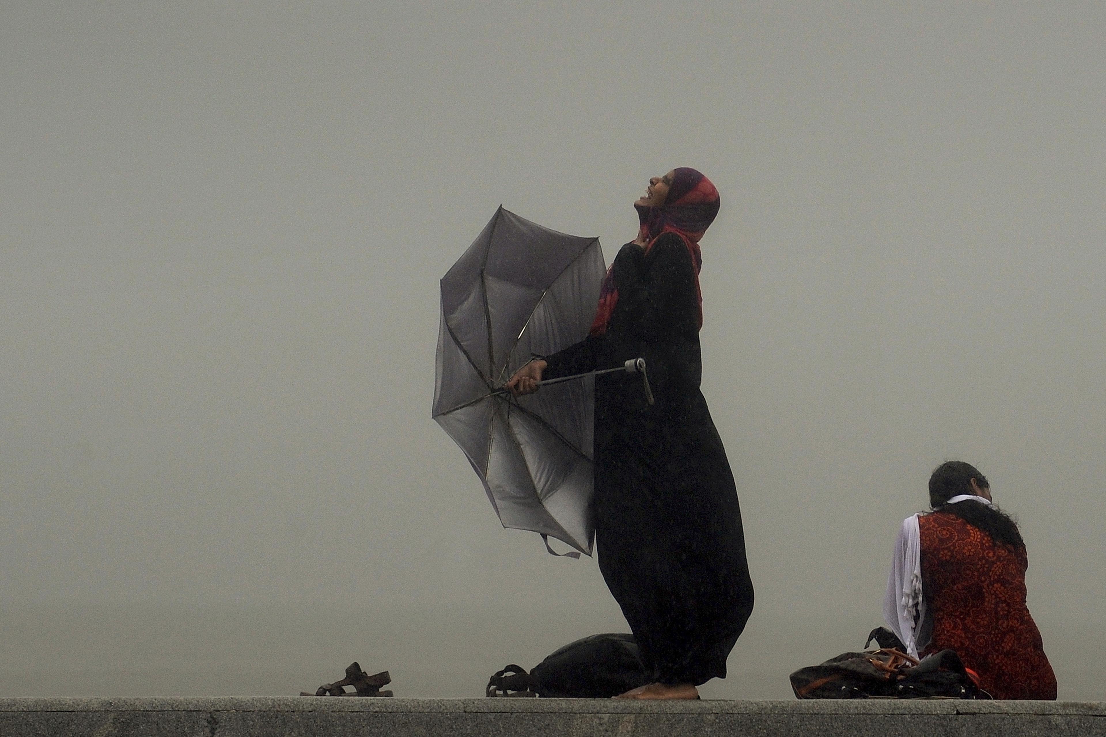 Photo of a woman in the rain holding a broken umbrella, smiling upwards; another sits nearby with a foggy background.