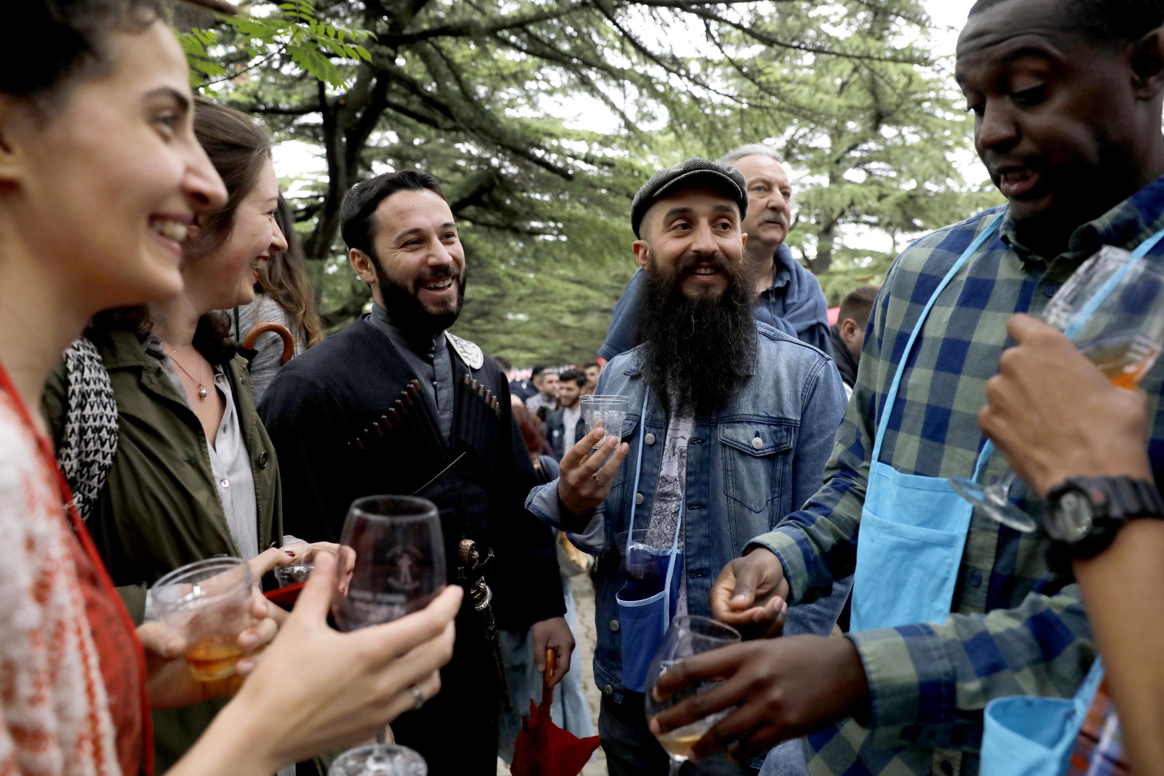 Photo of a diverse group of people laughing and holding drinks outdoors, surrounded by trees.