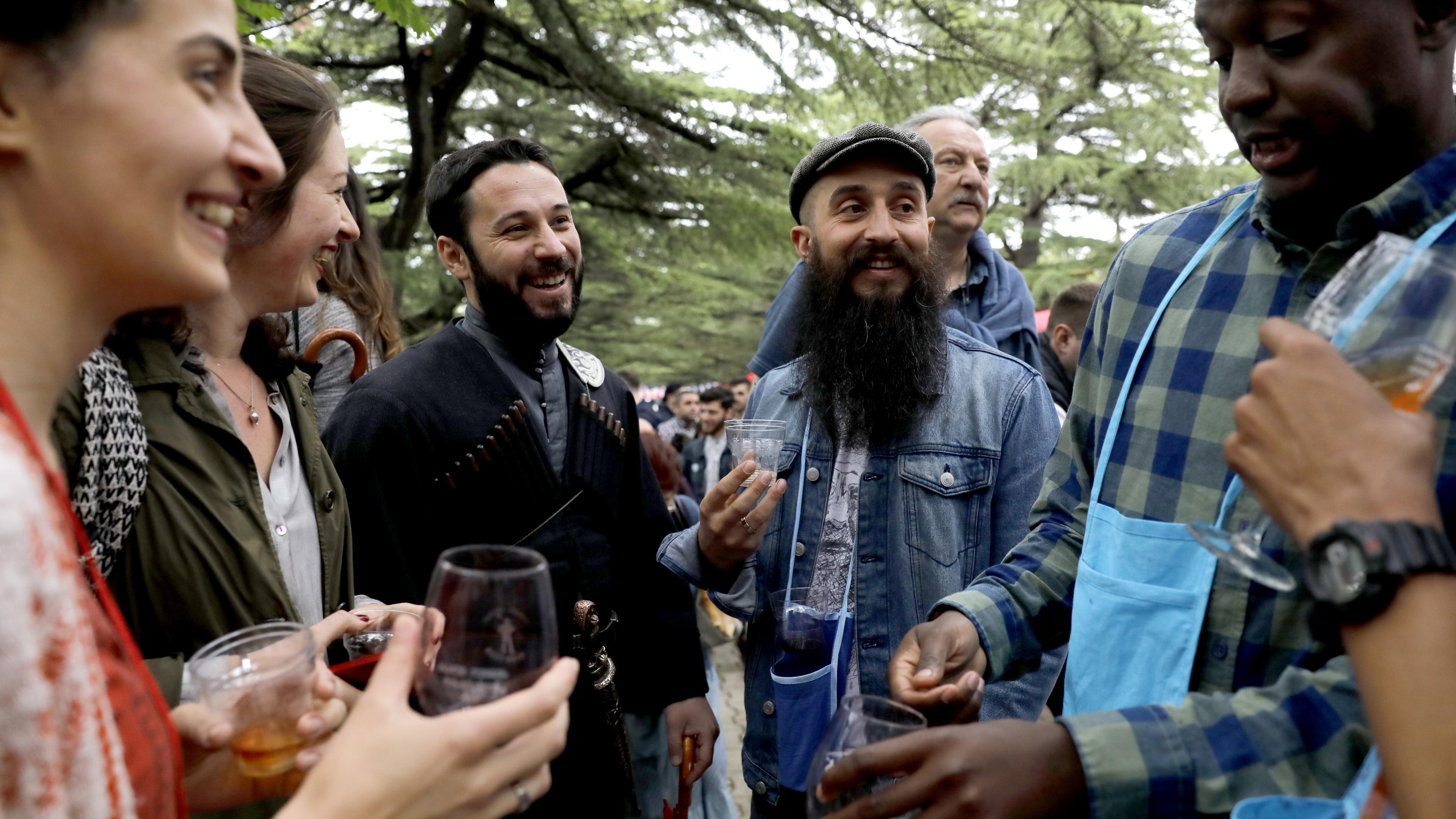 Photo of a diverse group of people laughing and holding drinks outdoors, surrounded by trees.