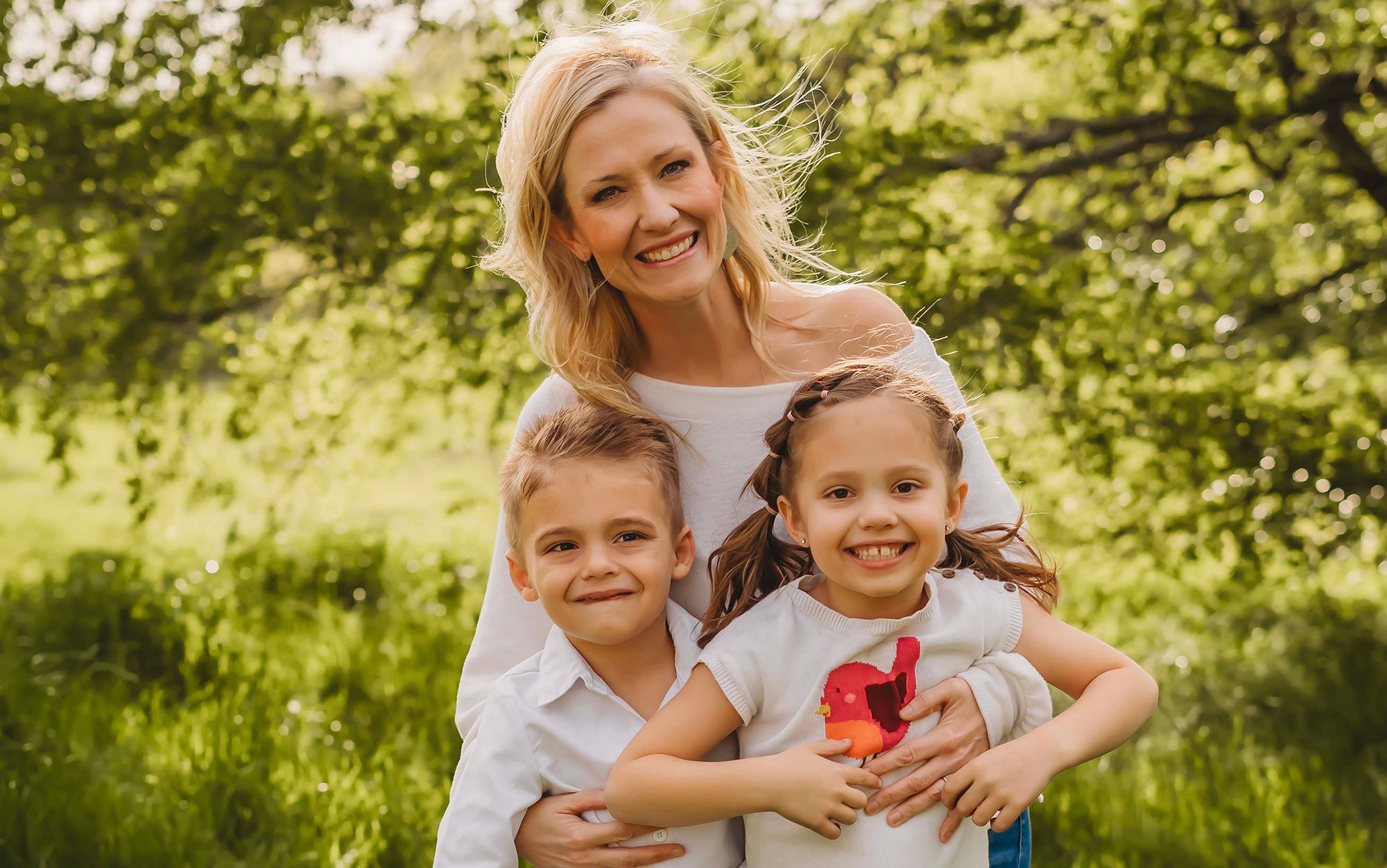 A smiling woman with two children in a sunny park, lush green trees in the background.