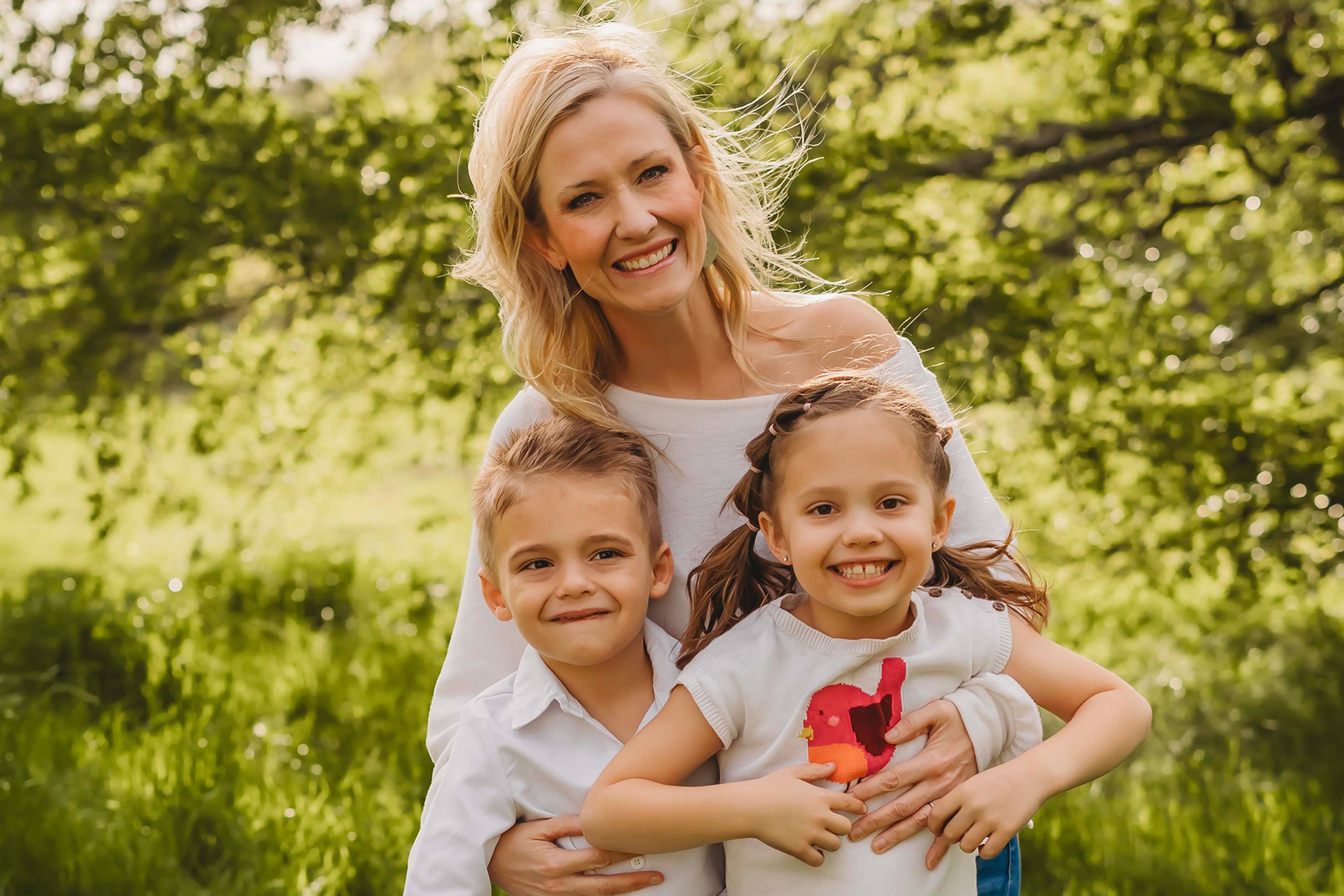 A smiling woman with two children in a sunny park, lush green trees in the background.