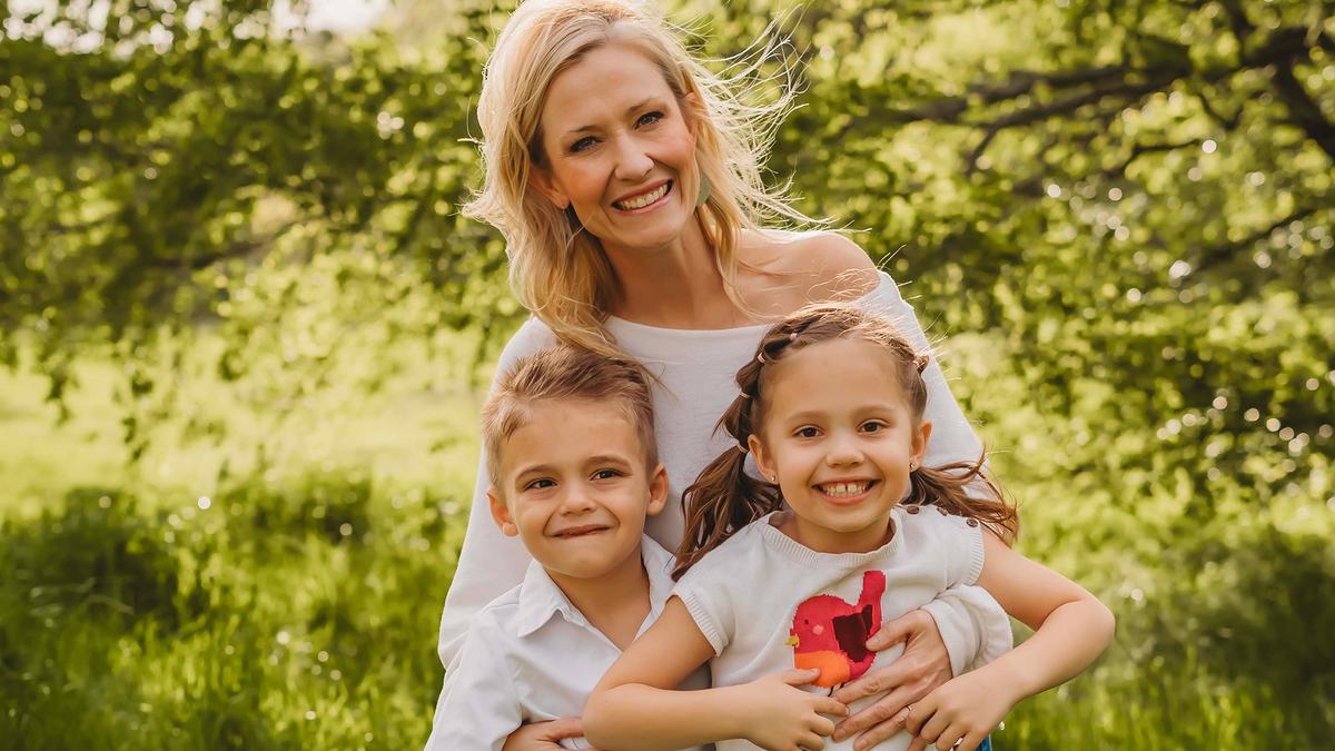 A smiling woman with two children in a sunny park, lush green trees in the background.