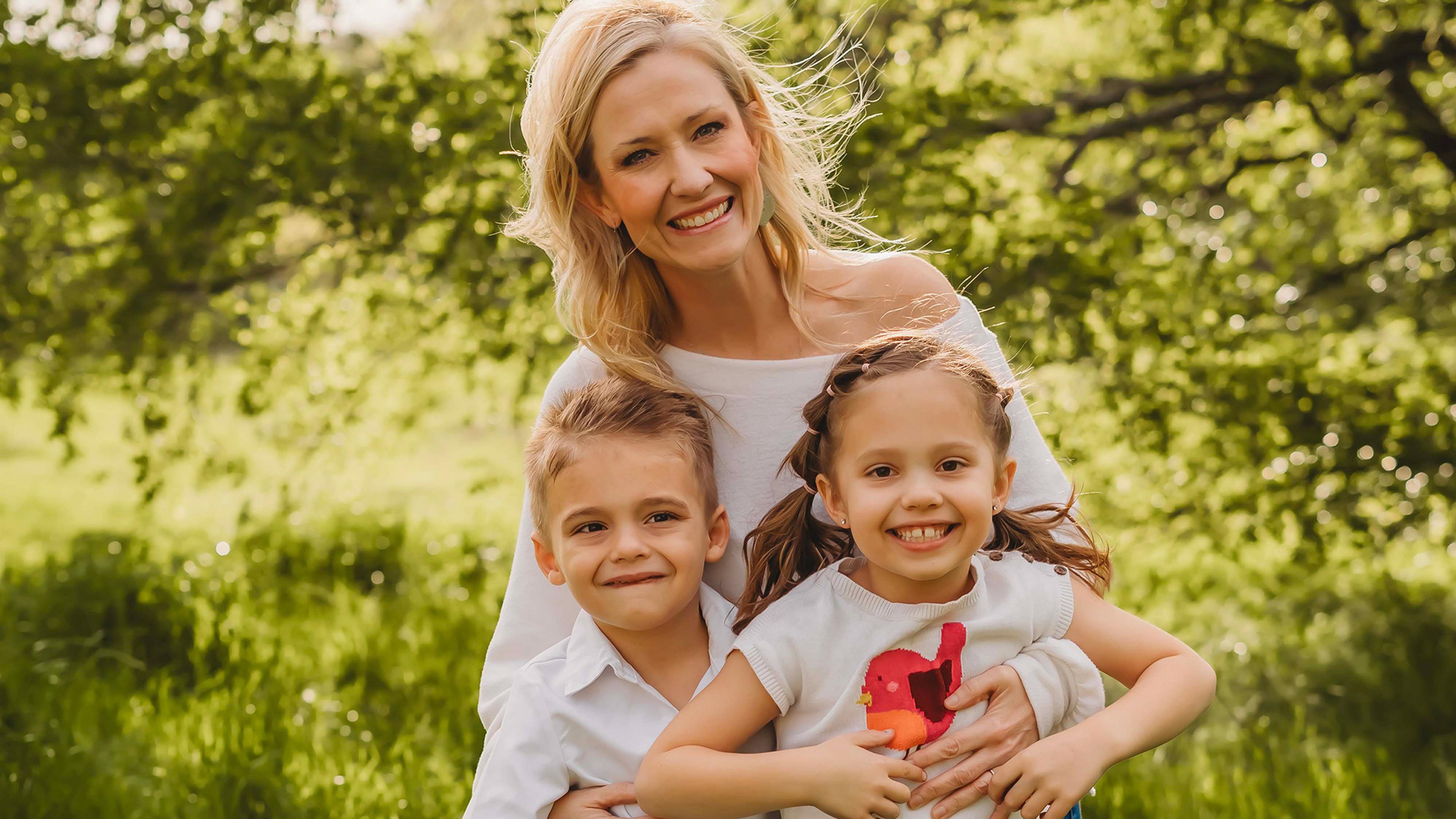 A smiling woman with two children in a sunny park, lush green trees in the background.