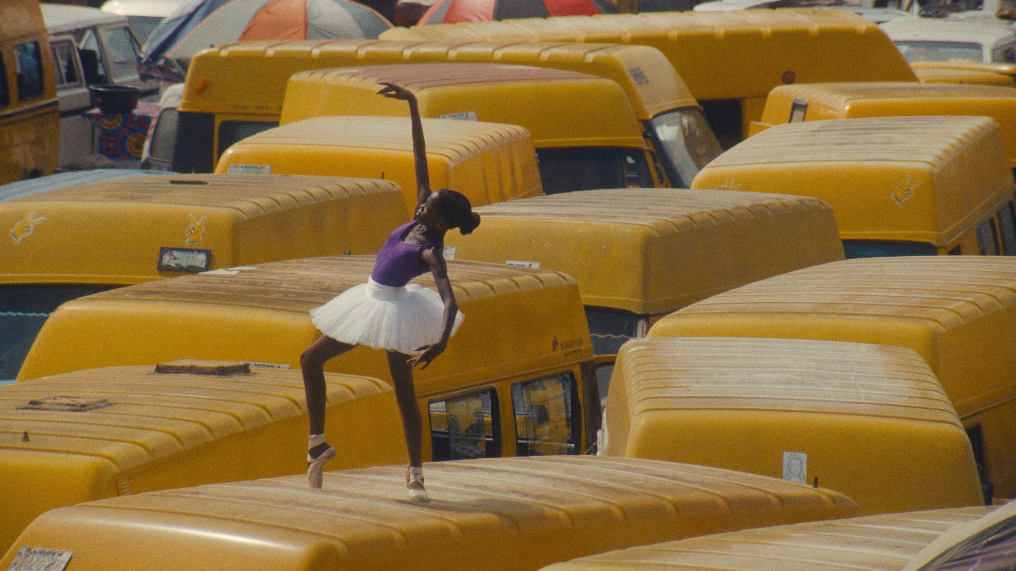 A Nigerian ballerina in a white tutu dancing atop yellow minibuses in a crowded urban setting.