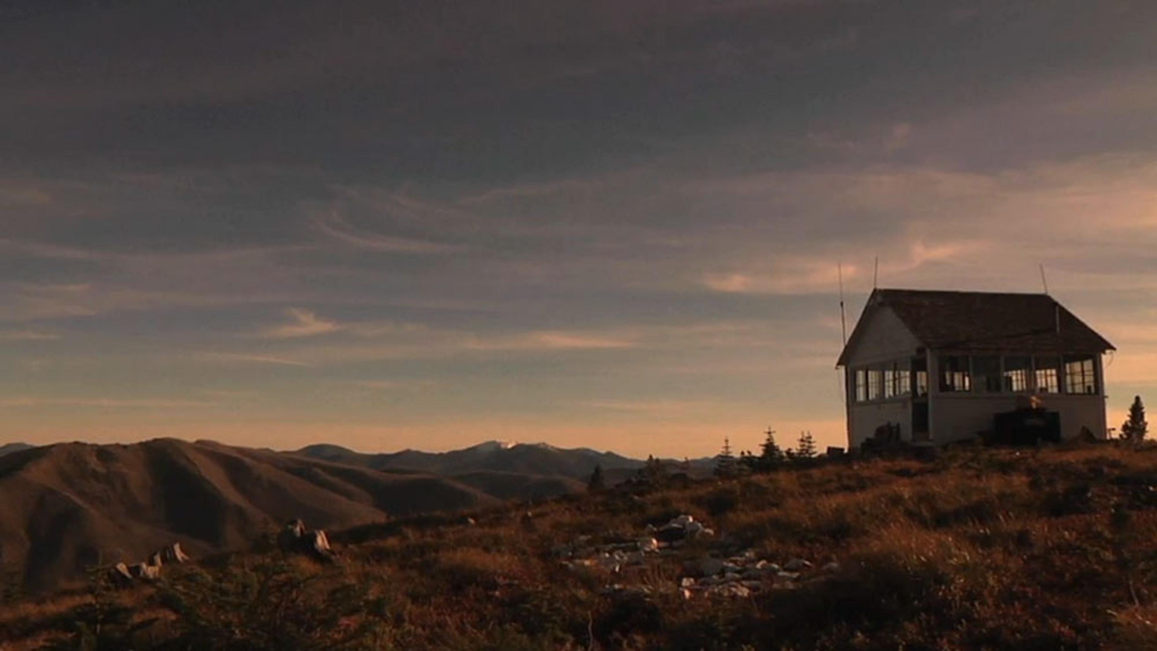 A solitary house on a hill during sunset with mountains and a cloudy sky in the background.