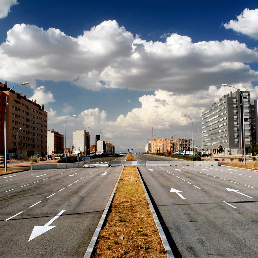 Wide, empty urban road flanked by modern apartment buildings under a partly cloudy sky.