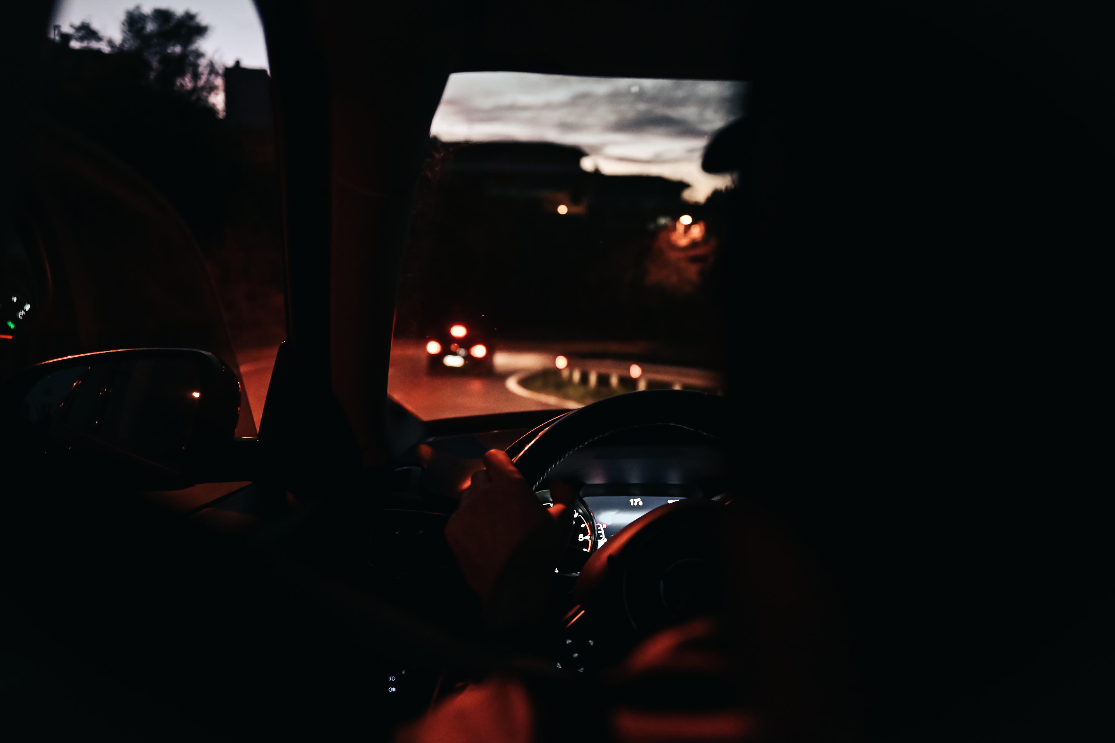 Photo of a person driving at night with dashboard lights on and a road visible ahead through the car windscreen.