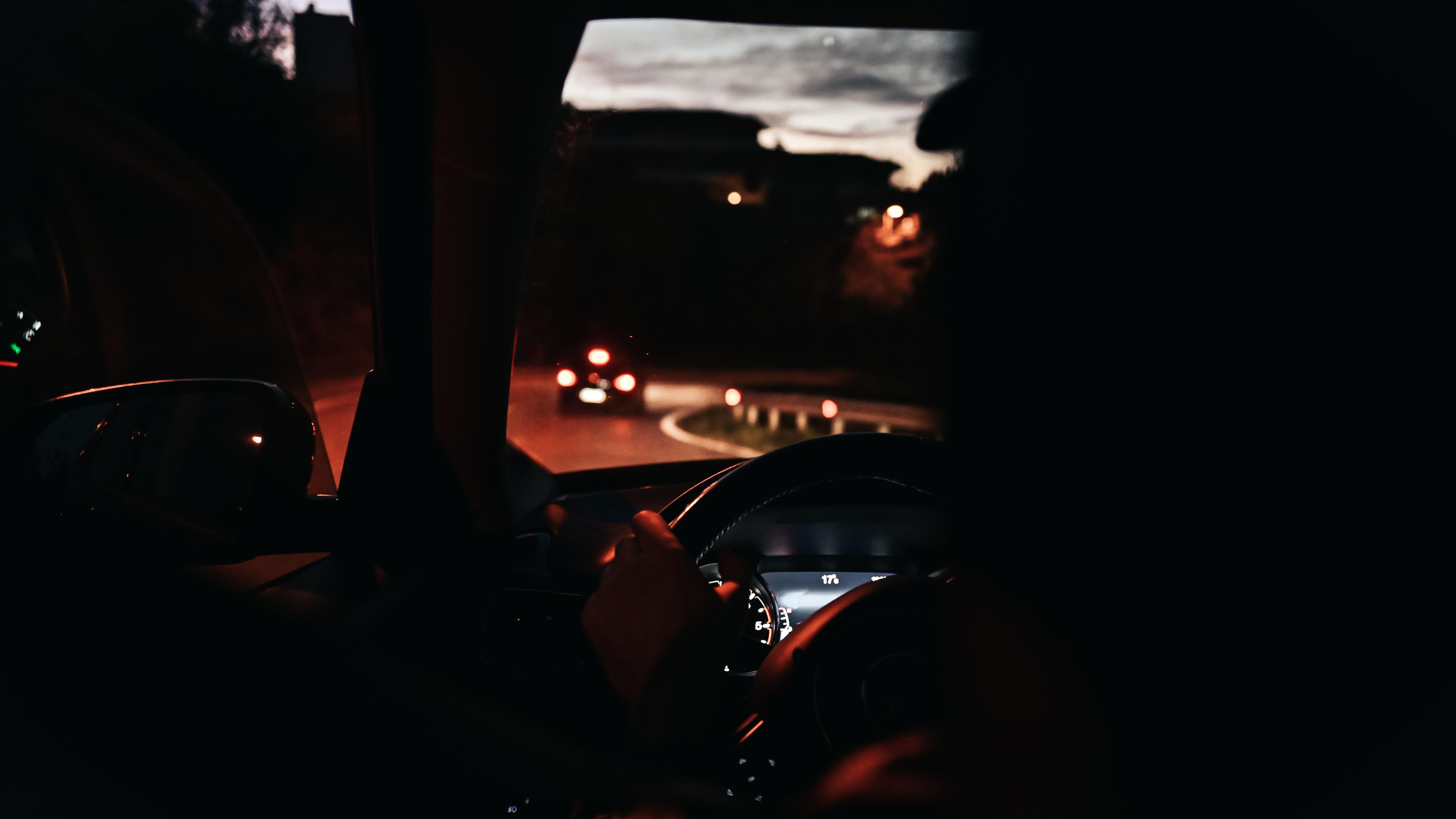 Photo of a person driving at night with dashboard lights on and a road visible ahead through the car windscreen.
