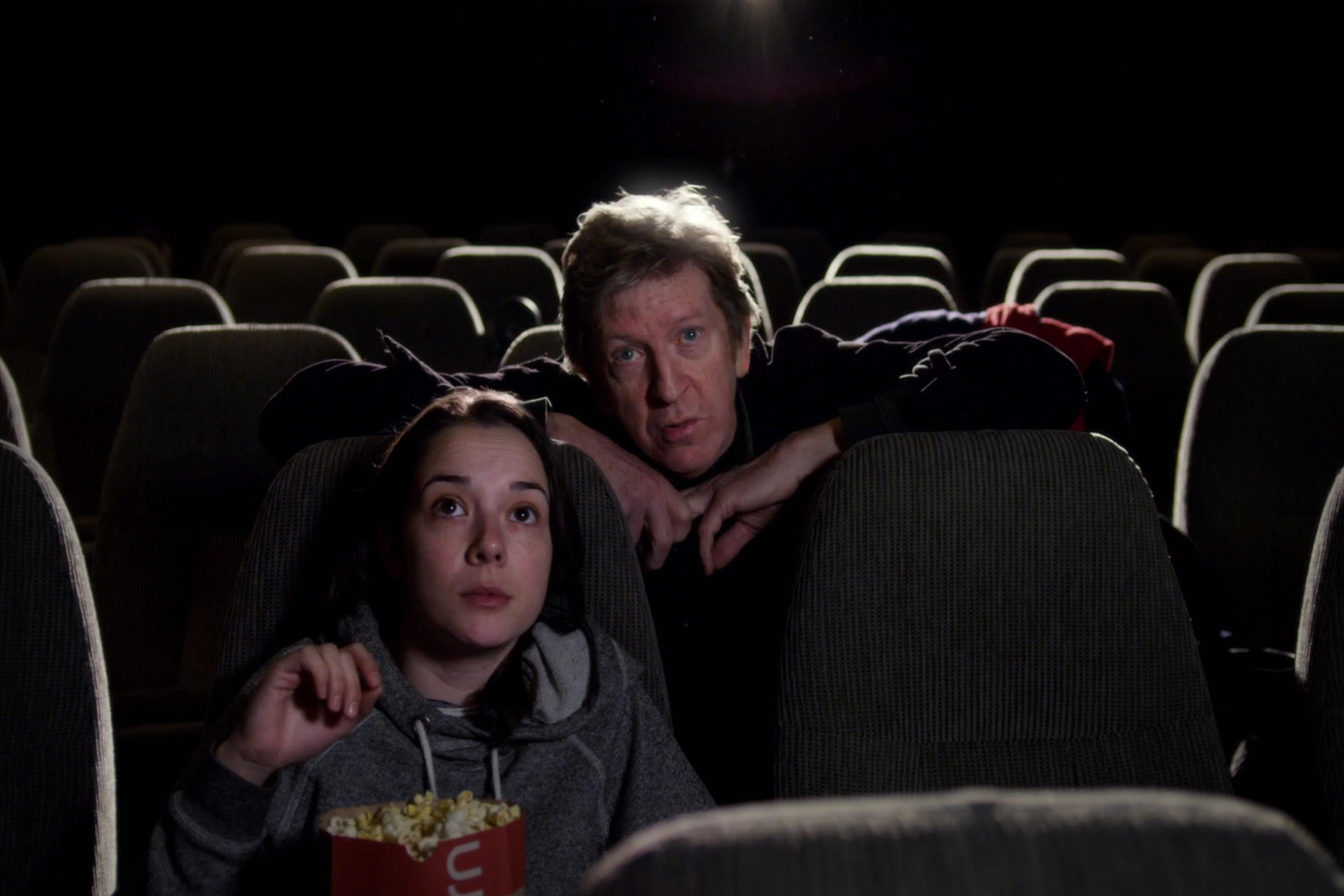 Two people watching a film in a dark, nearly empty cinema. One person is holding a popcorn box.