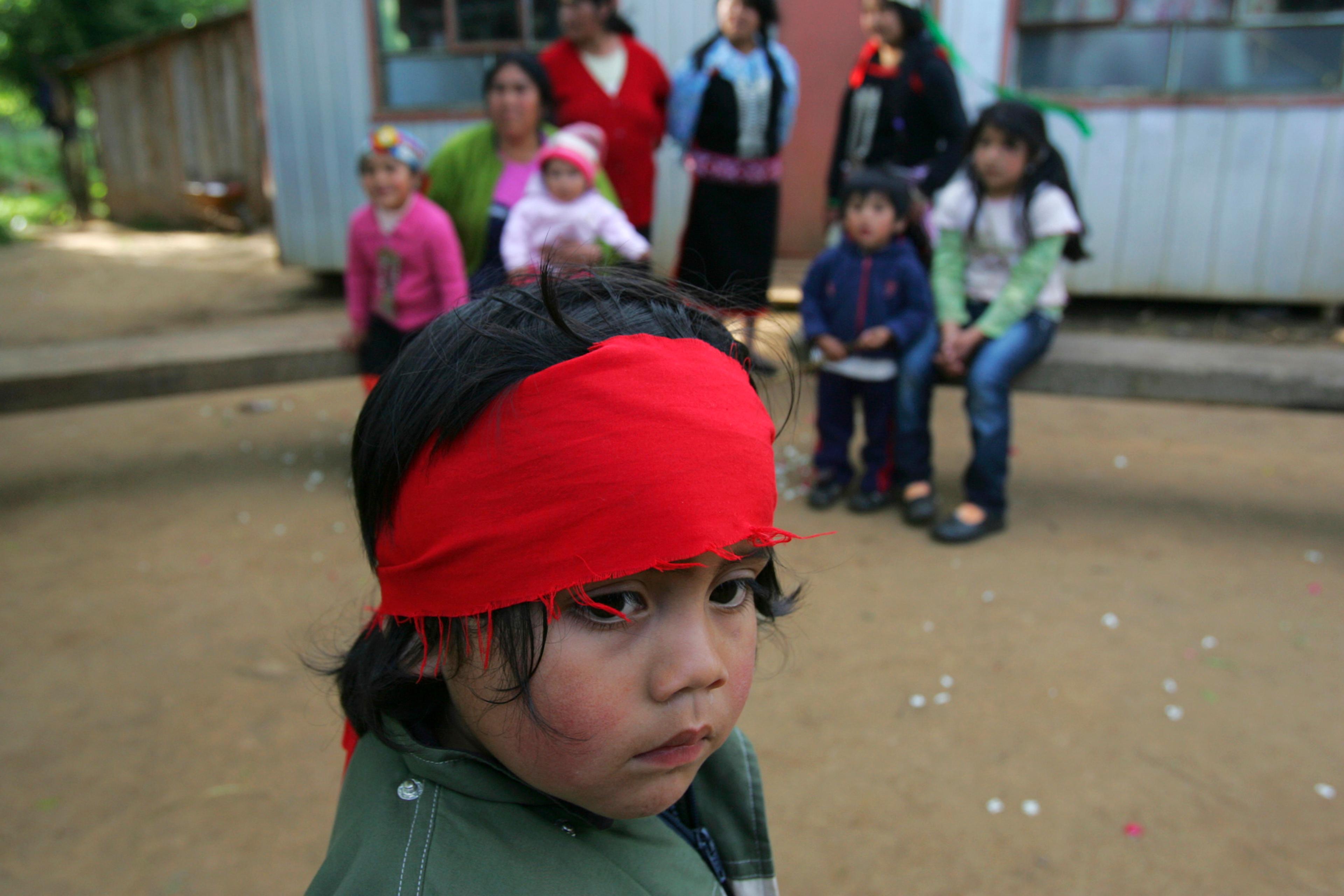 Photo of a child in a red headband in the foreground with other children and adults sitting outside a building in the background.