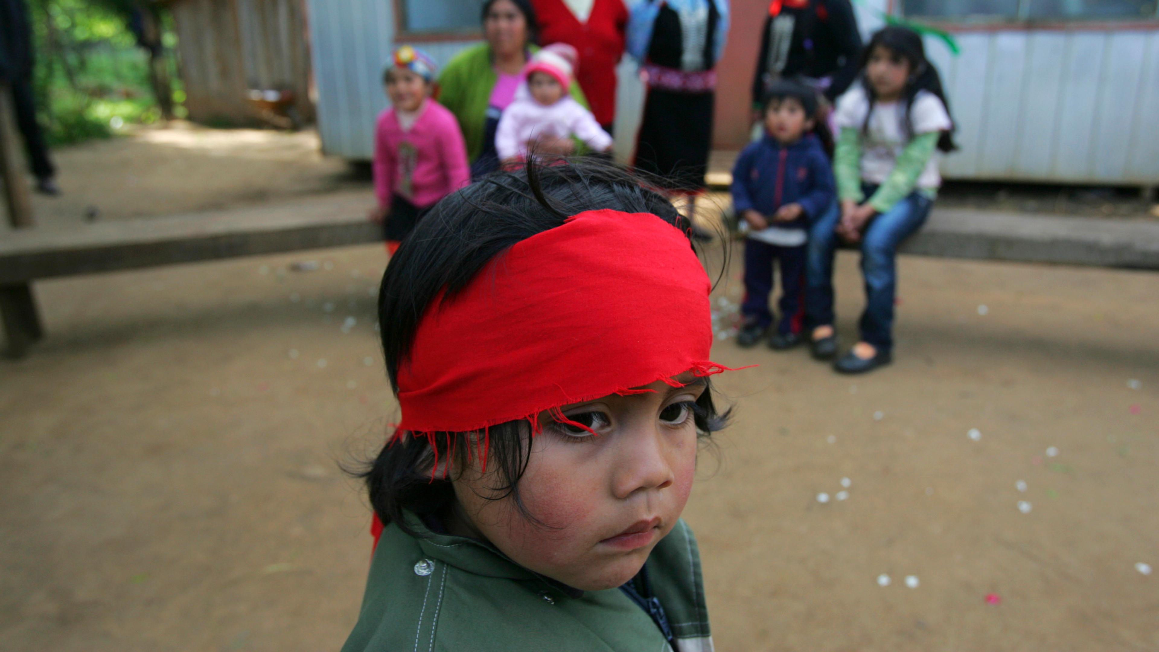 Photo of a child in a red headband in the foreground with other children and adults sitting outside a building in the background.