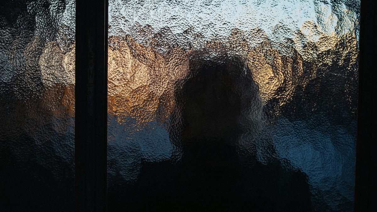 Silhouette of a person behind textured glass with a blurred background of sky and trees at sunset, photograph.