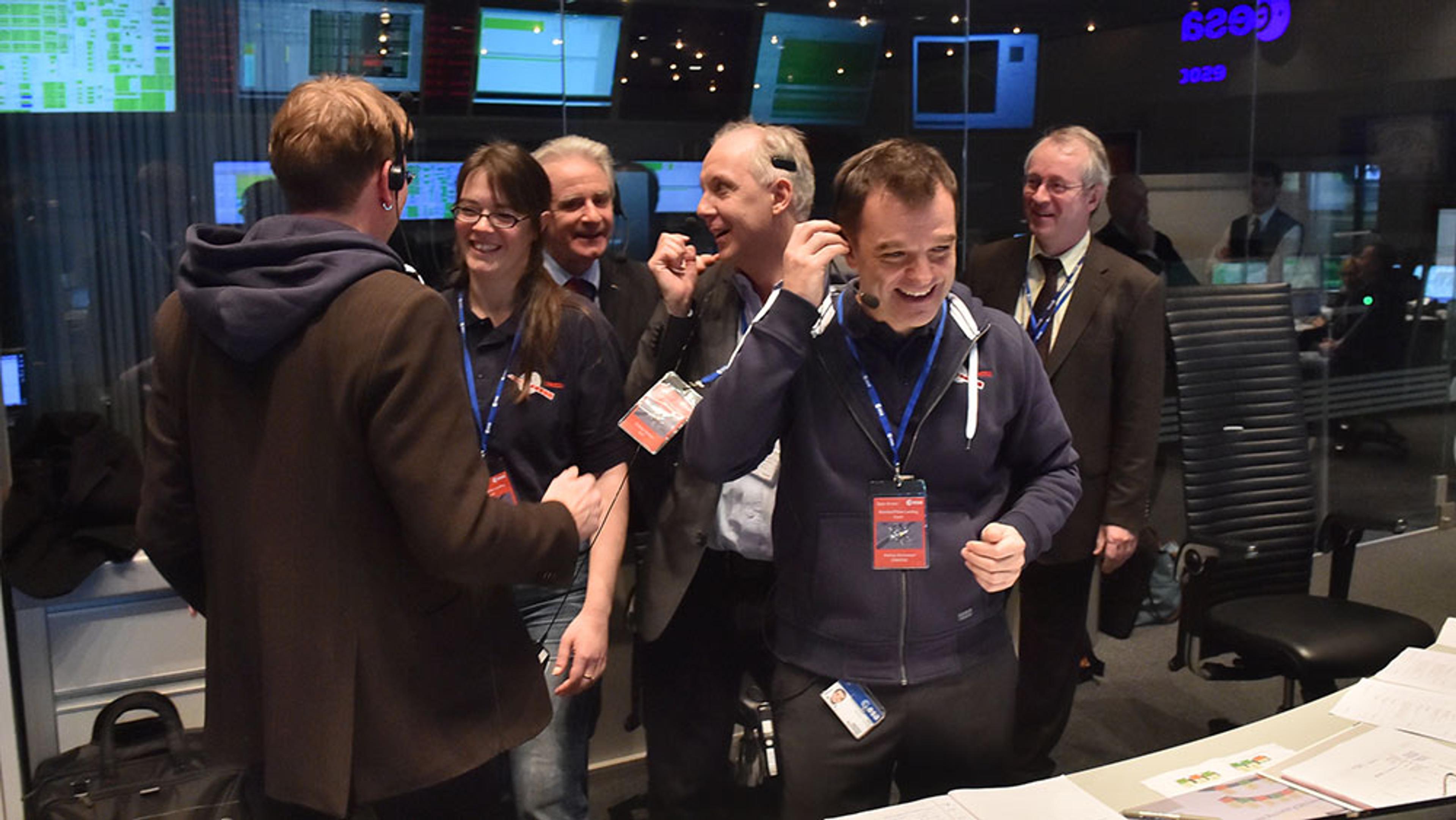 A group of people in an office celebrating with smiles and high-fives, standing near computer monitors.