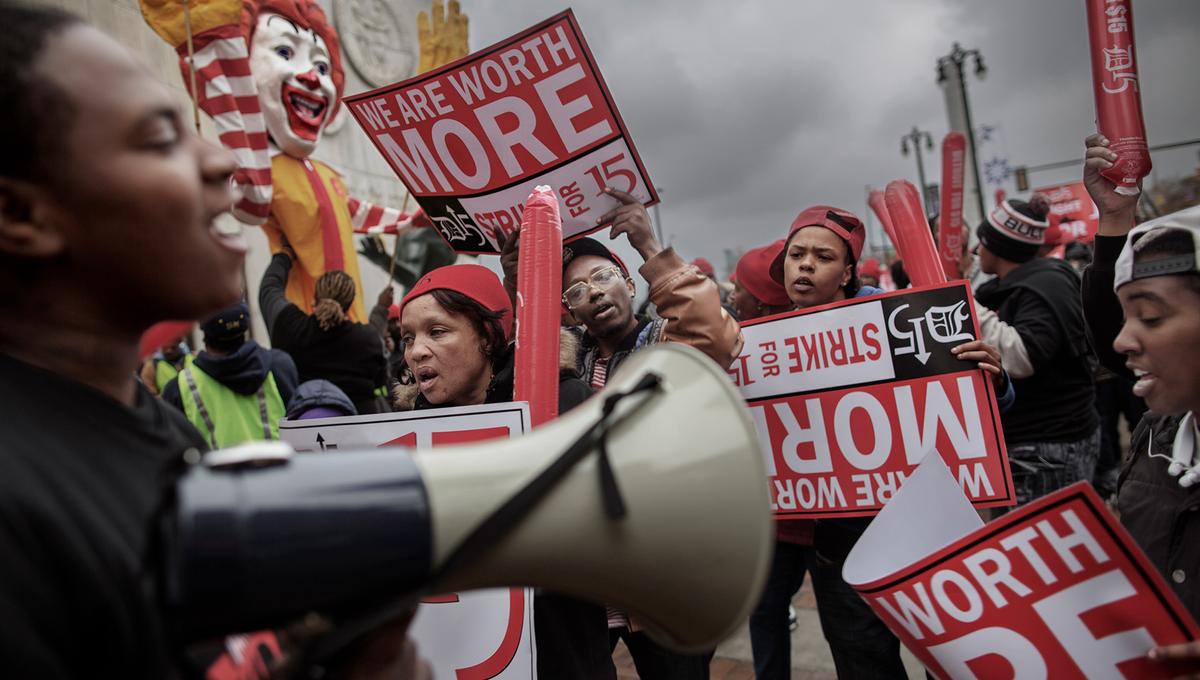 A protest with people holding signs that read ‘We are worth more’ and ‘Strike for 15’; large Ronald McDonald figure in the background