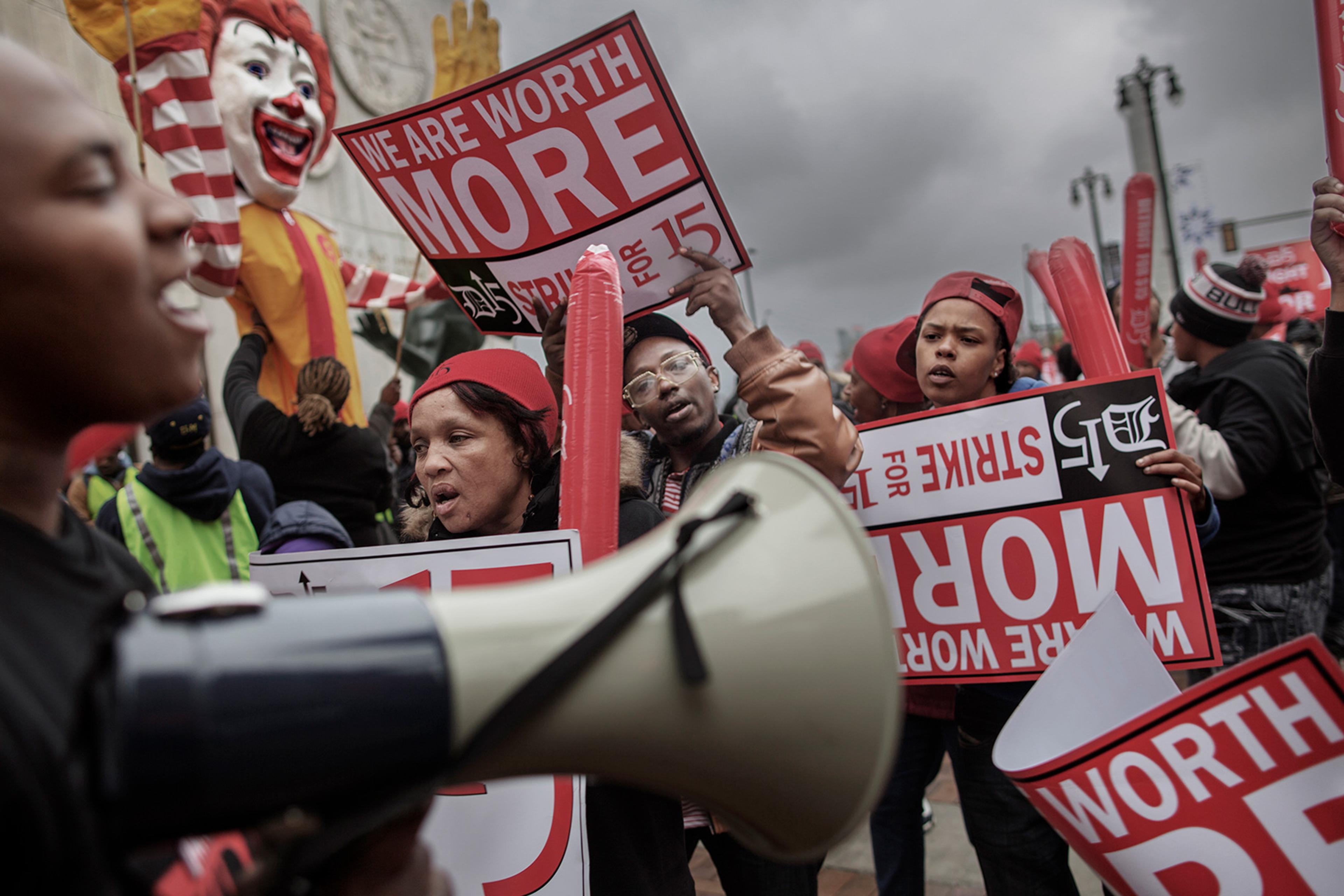 A protest with people holding signs that read ‘We are worth more’ and ‘Strike for 15’; large Ronald McDonald figure in the background