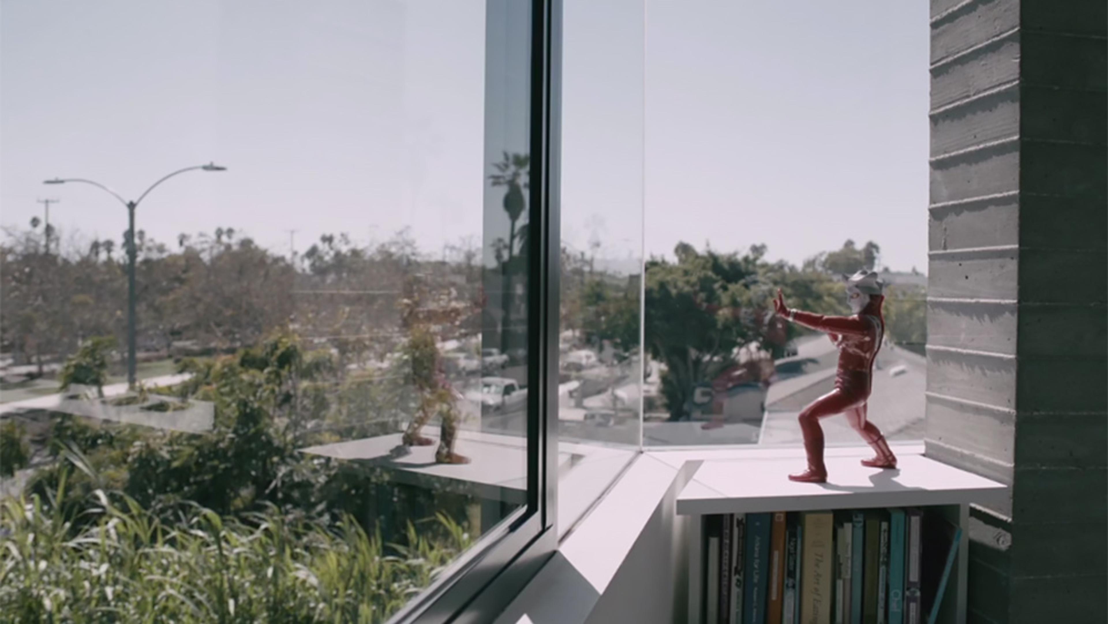 A small red figurine on a white shelf by a window overlooking a street with trees and buildings in the background.