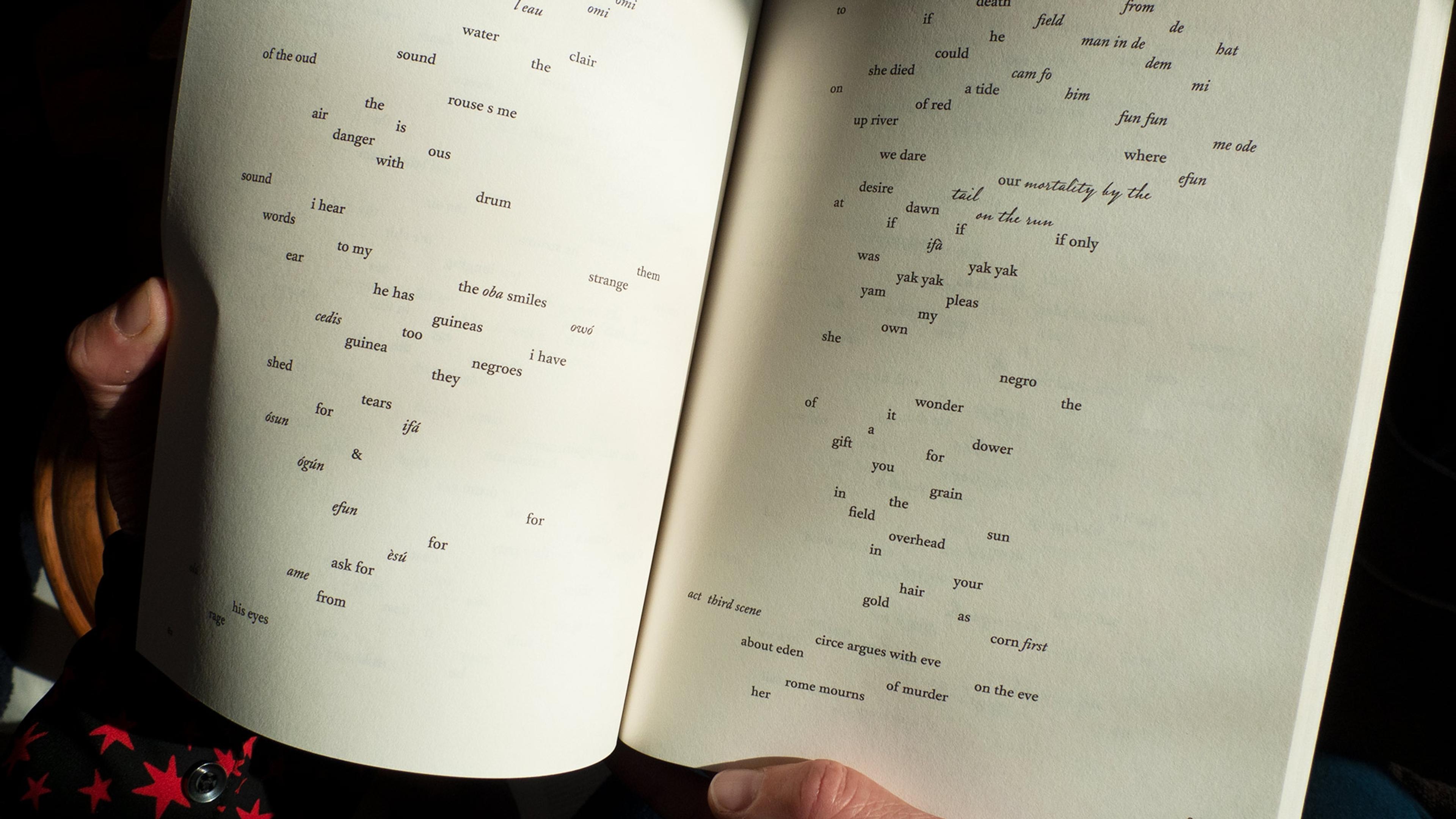 Photo of an open book with scattered text, held by hands. The text appears as a poetic or artistic composition.