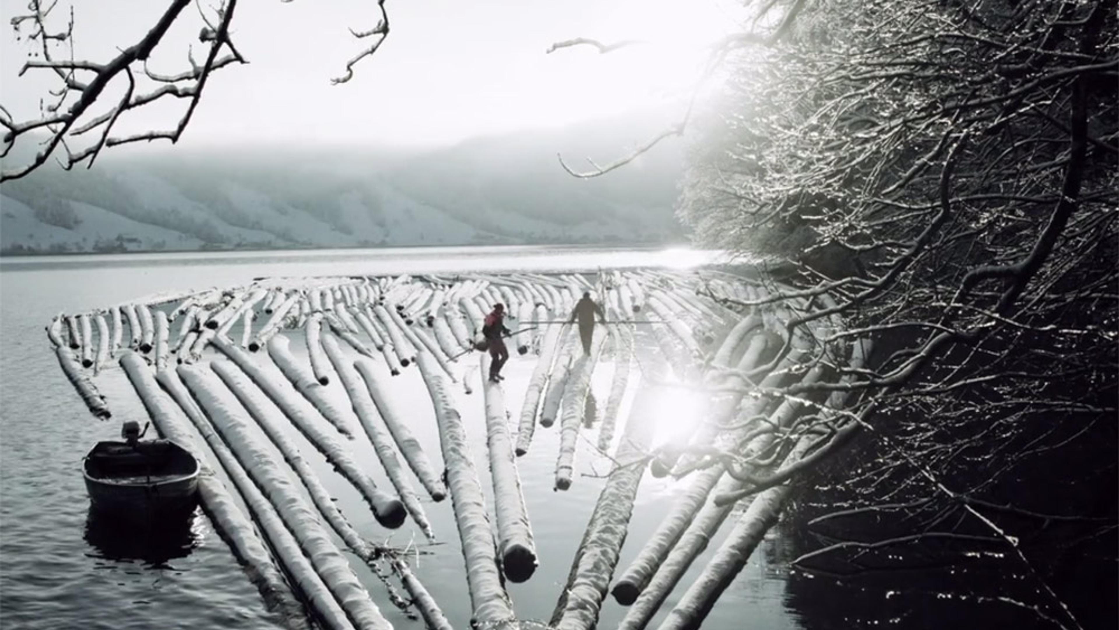 Photo of two people walking on snow-covered logs floating on a lake with a boat and snowy tree branches in the foreground.