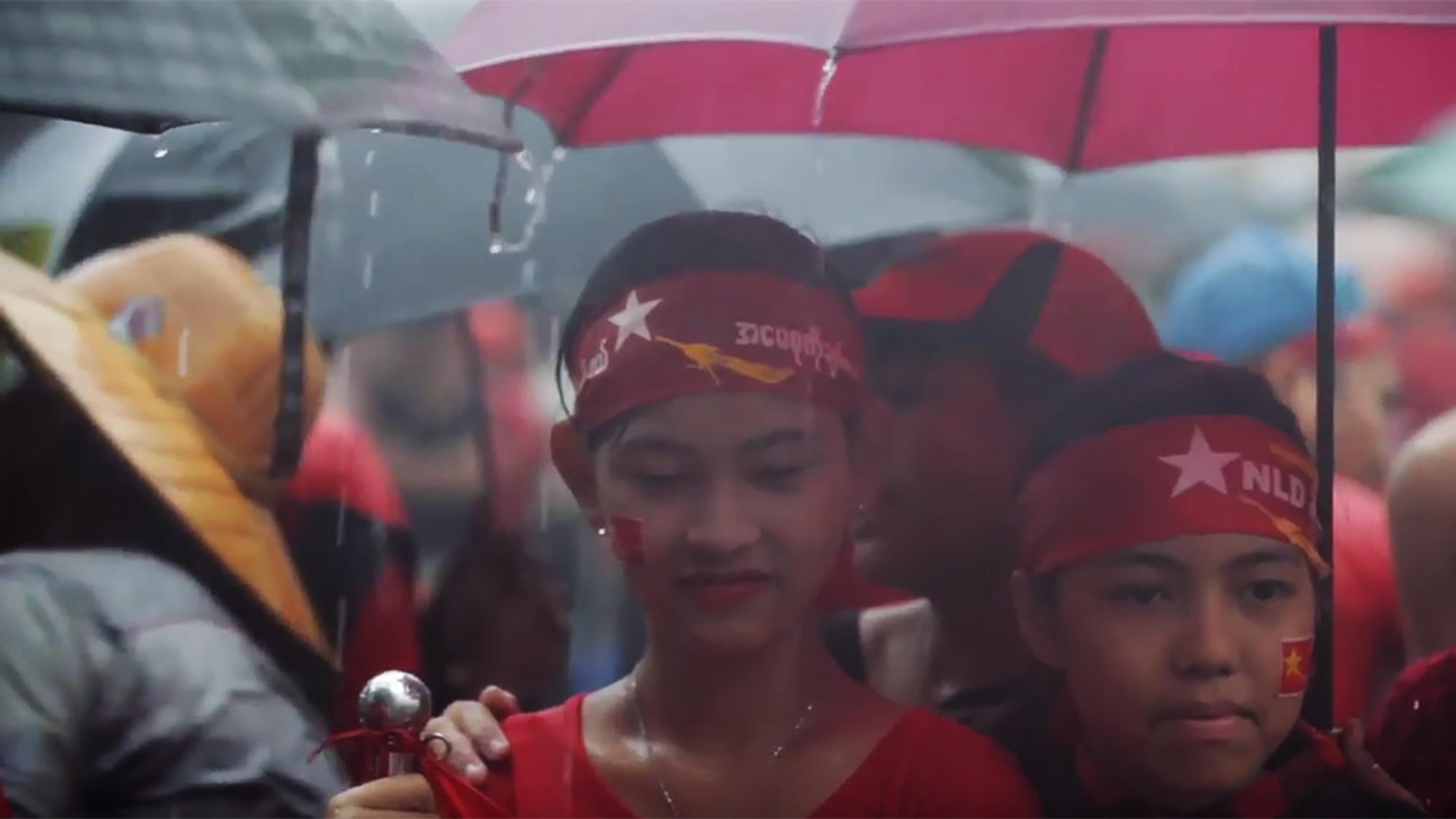 Two people in red headbands and shirts under an umbrella in the rain at a political demonstration in Myanmar.