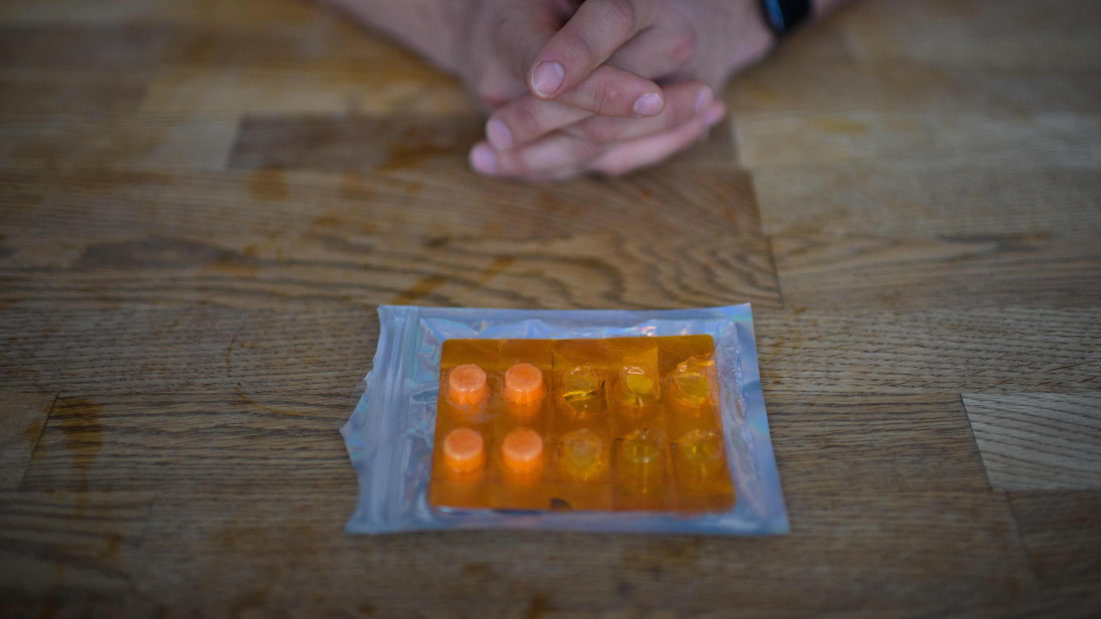 Photo of hands clasped on a wooden table with a blister pack of orange pills in front.