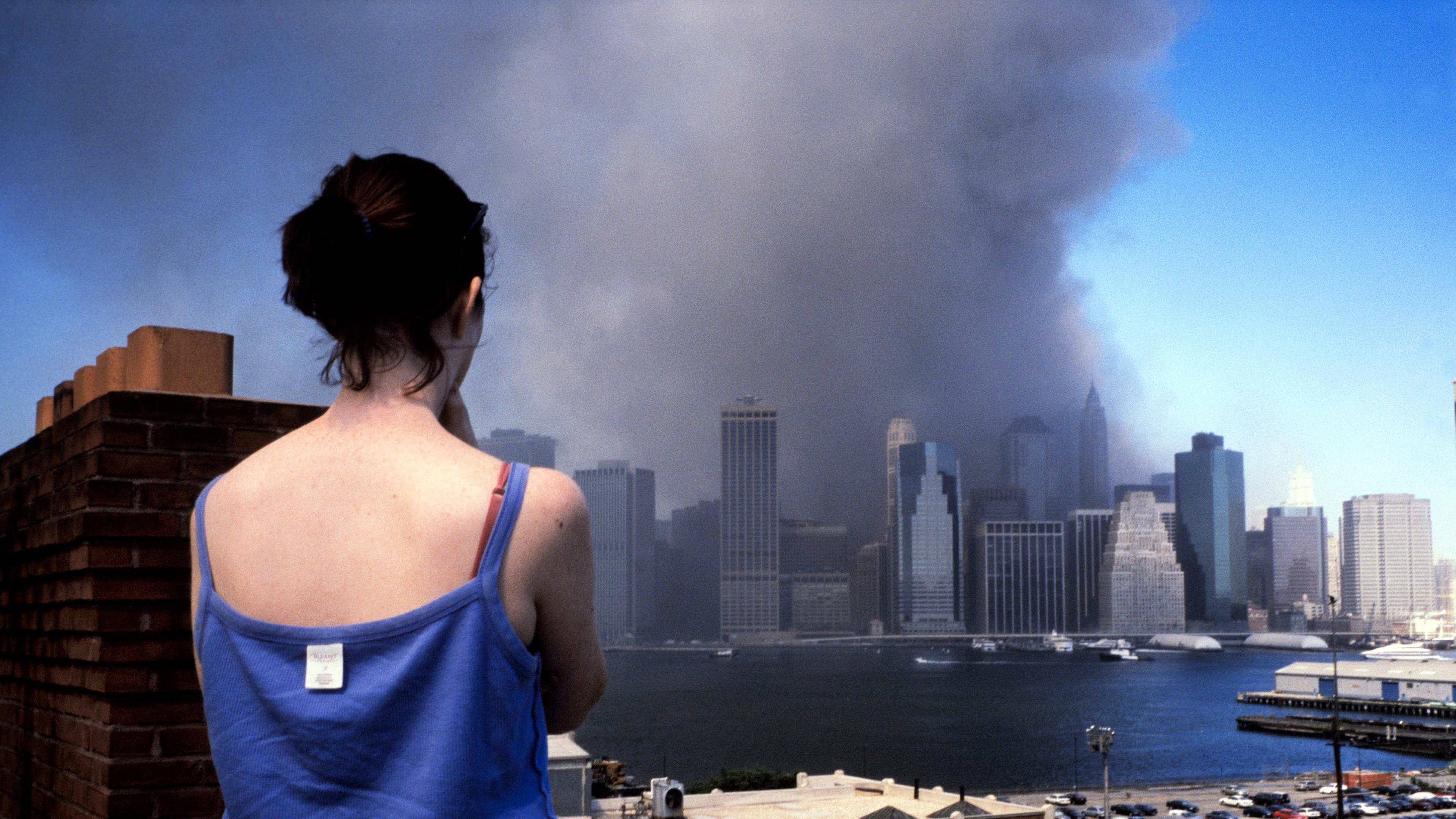 Photo of a woman in a blue top watching smoke rise from skyscrapers across a river under a clear blue sky.