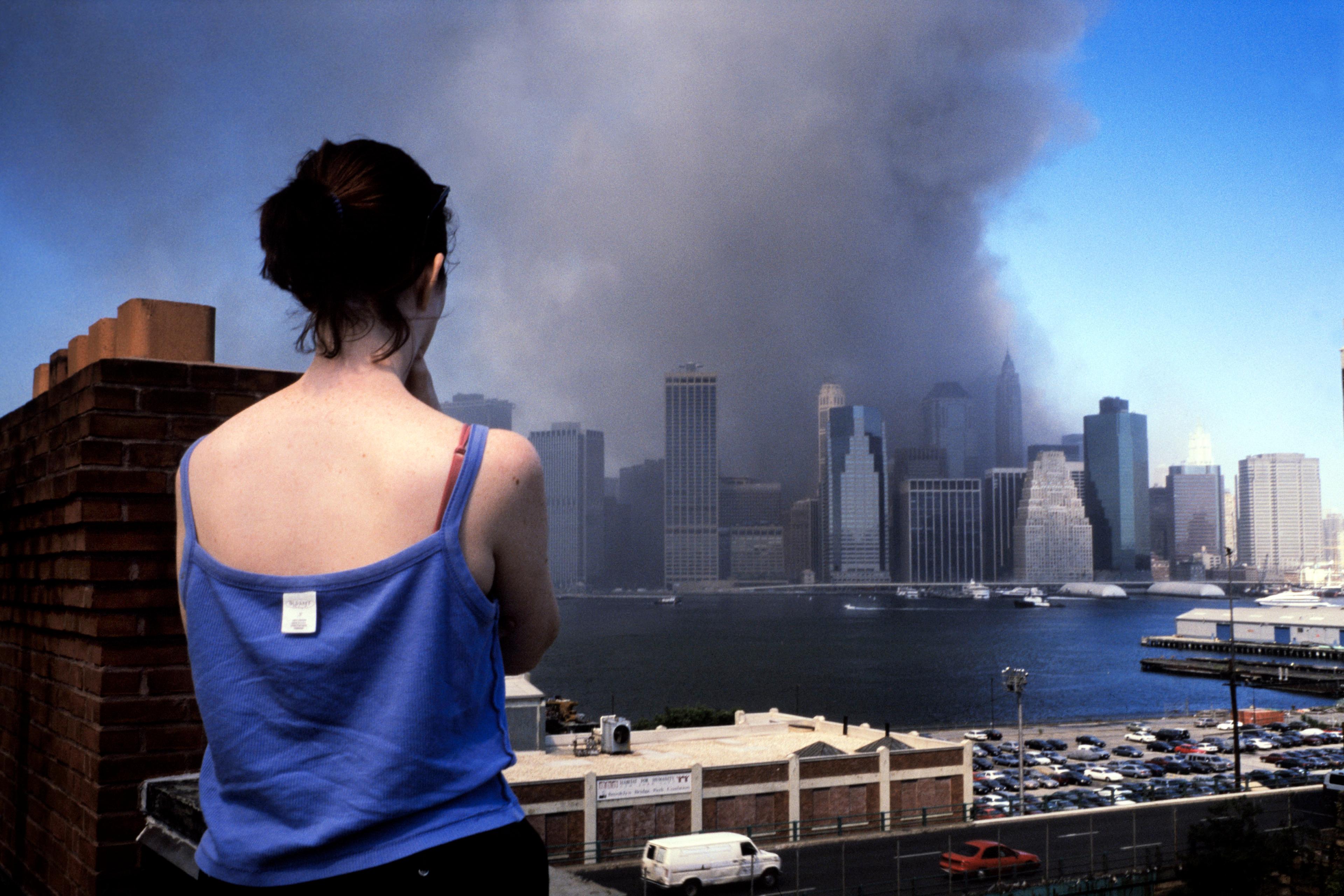 Photo of a woman in a blue top watching smoke rise from skyscrapers across a river under a clear blue sky.