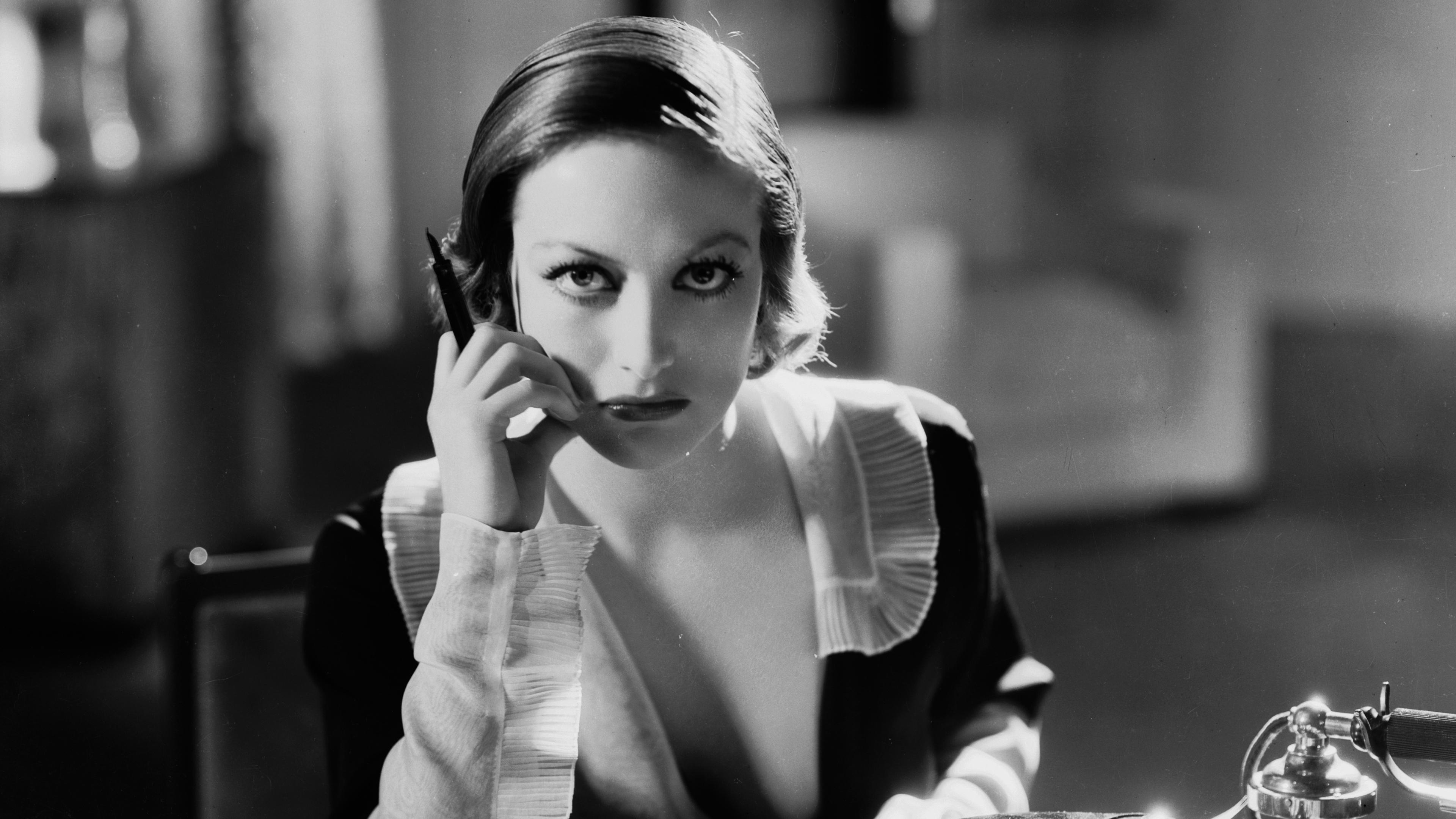 Black and white photo of a woman at a desk with a typewriter and a phone, deep in thought, holding a pen to her cheek.