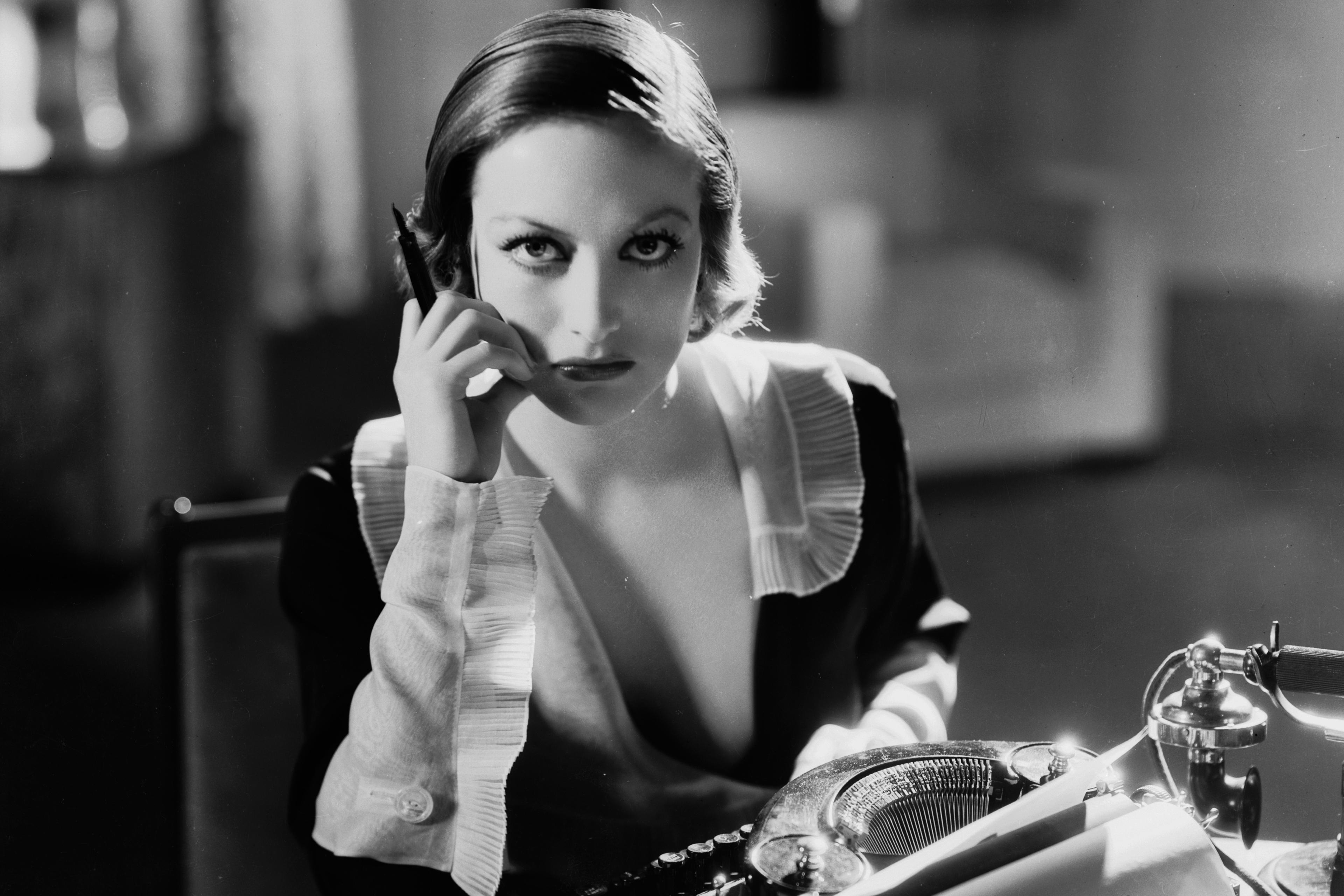 Black and white photo of a woman at a desk with a typewriter and a phone, deep in thought, holding a pen to her cheek.