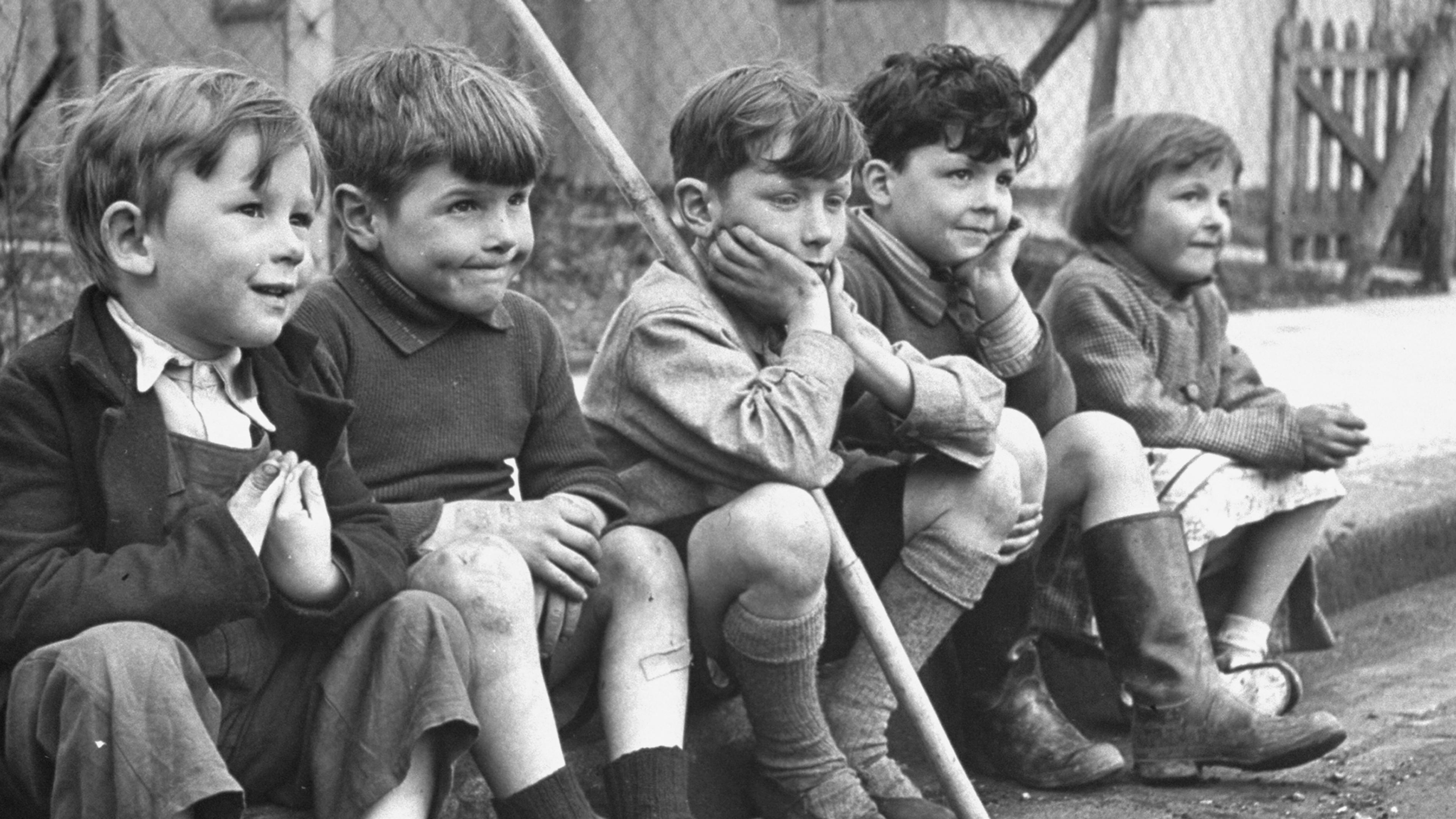 Black and white photo of five children sitting on a kerbside in front of a house, with playful expressions.