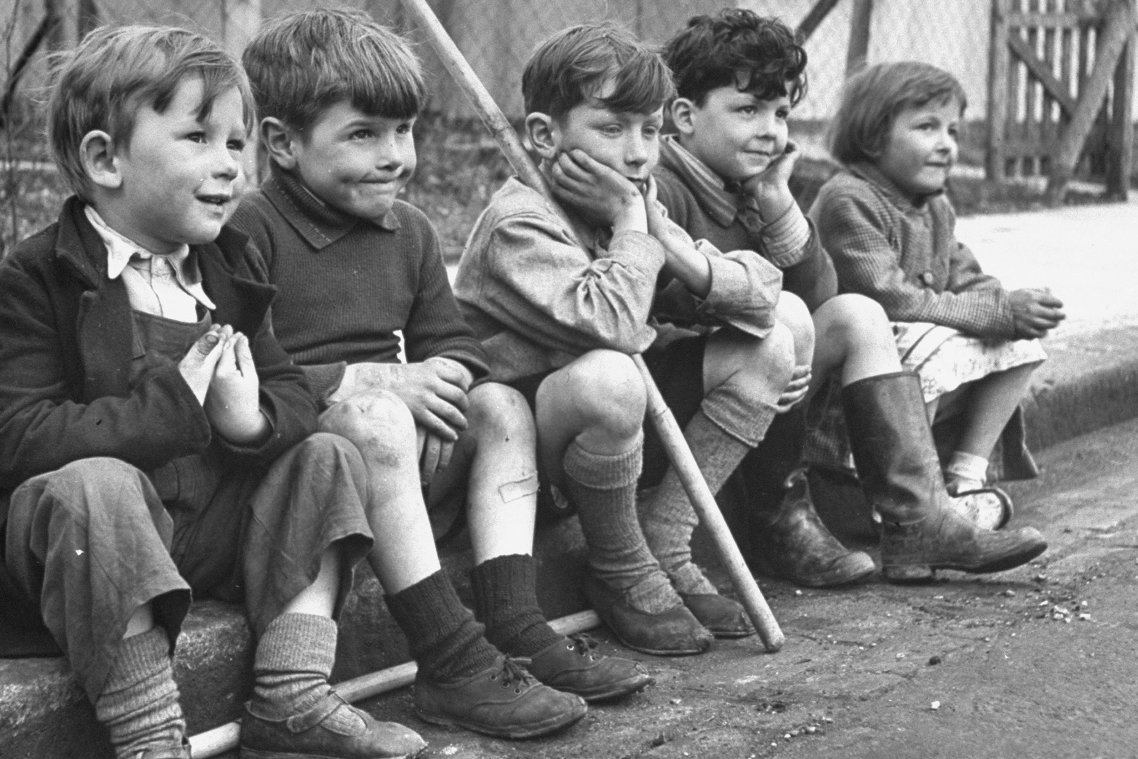 Black and white photo of five children sitting on a kerbside in front of a house, with playful expressions.