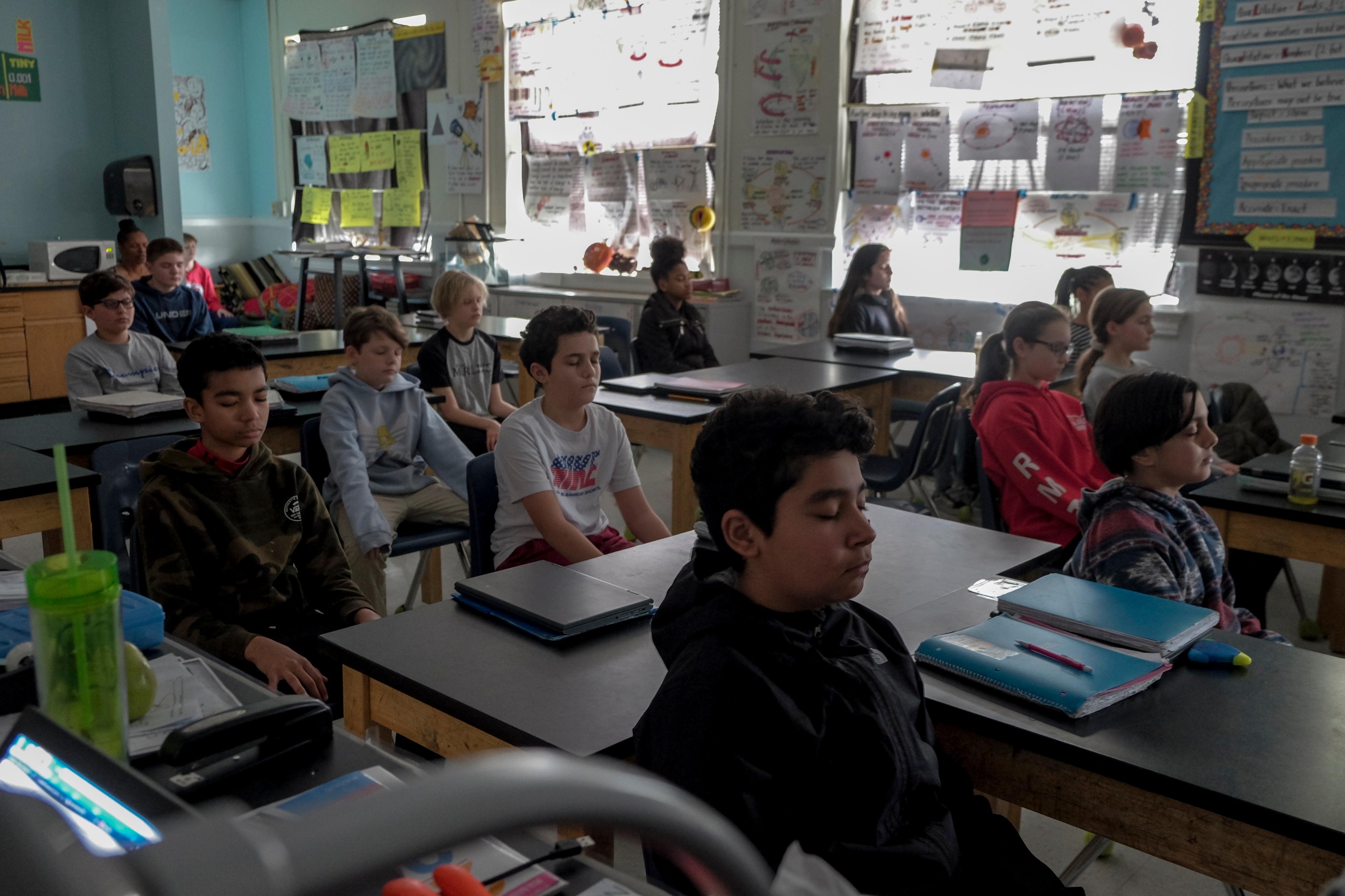 Photo of a classroom with students meditating at their desks, surrounded by posters and bright natural light.