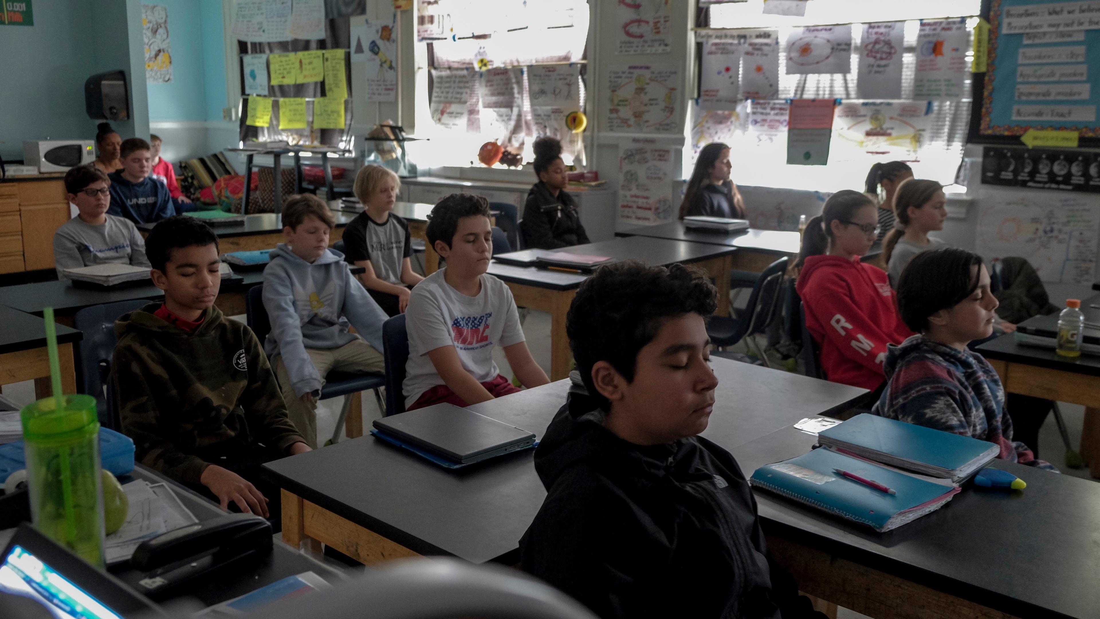 Photo of a classroom with students meditating at their desks, surrounded by posters and bright natural light.