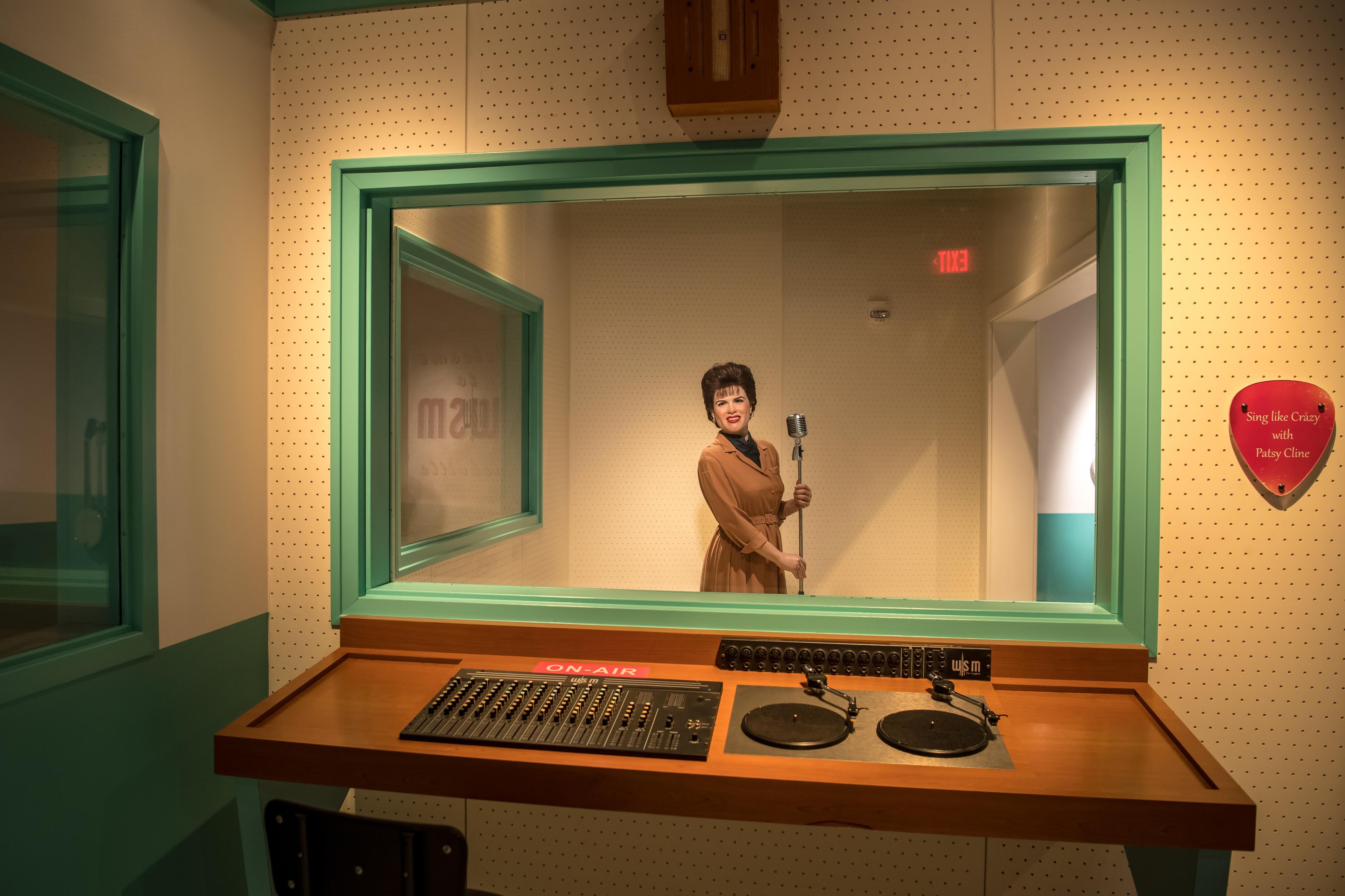 Photo of a vintage recording studio with a mannequin holding a microphone seen through a glass window from the control room.