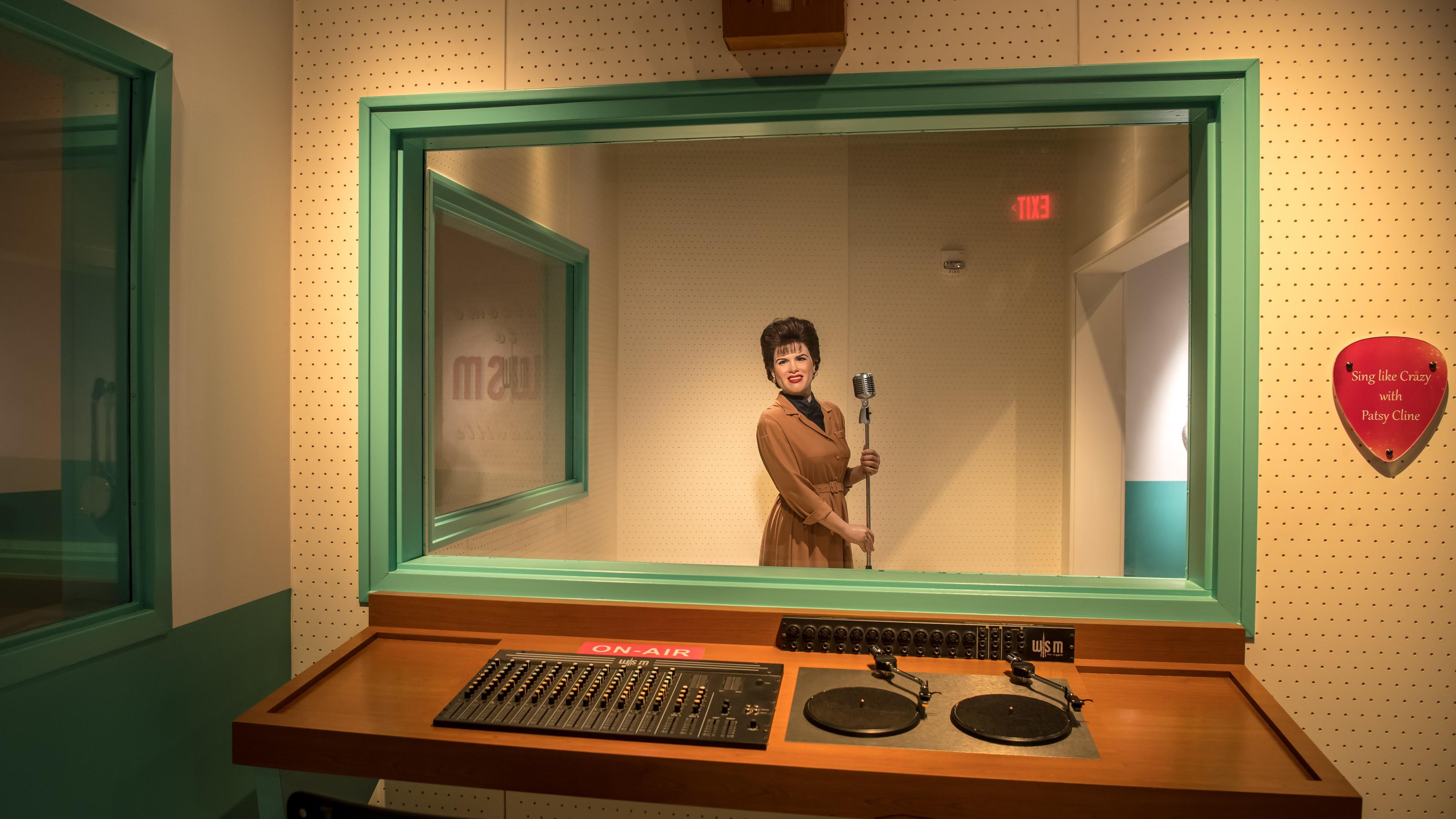 Photo of a vintage recording studio with a mannequin holding a microphone seen through a glass window from the control room.