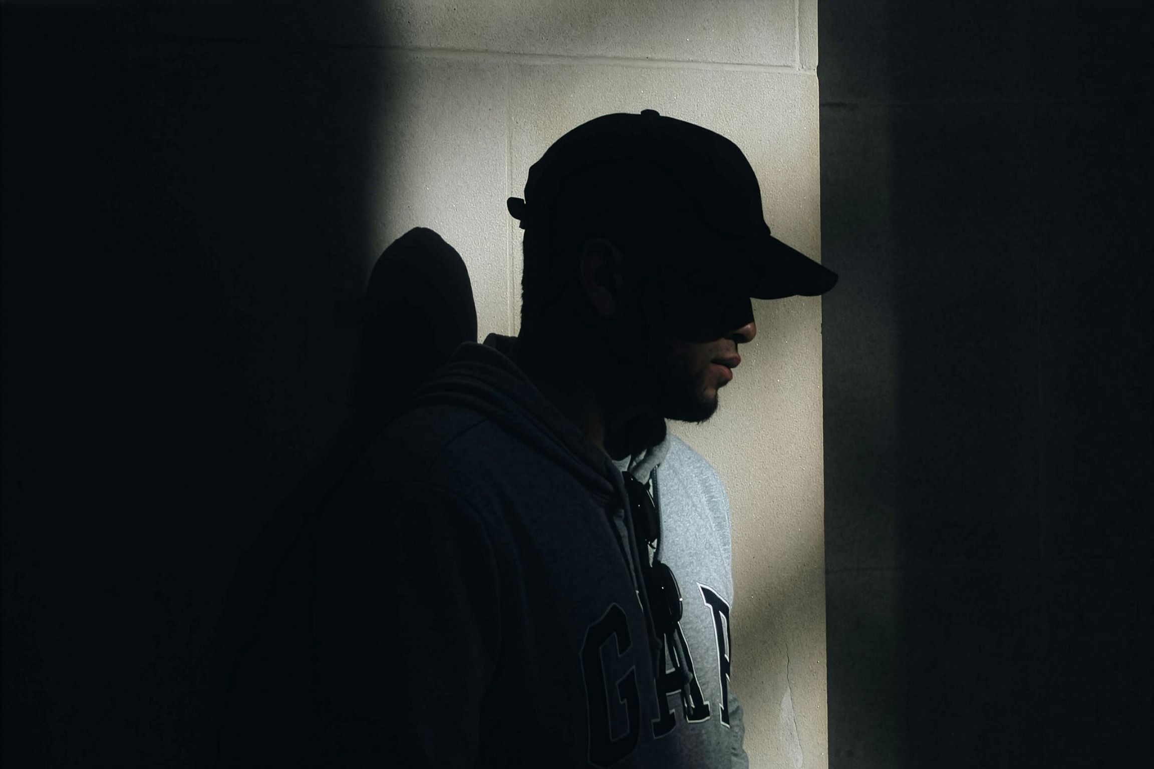 A Black man in a cap and hoodie standing in shadow against a lit wall.