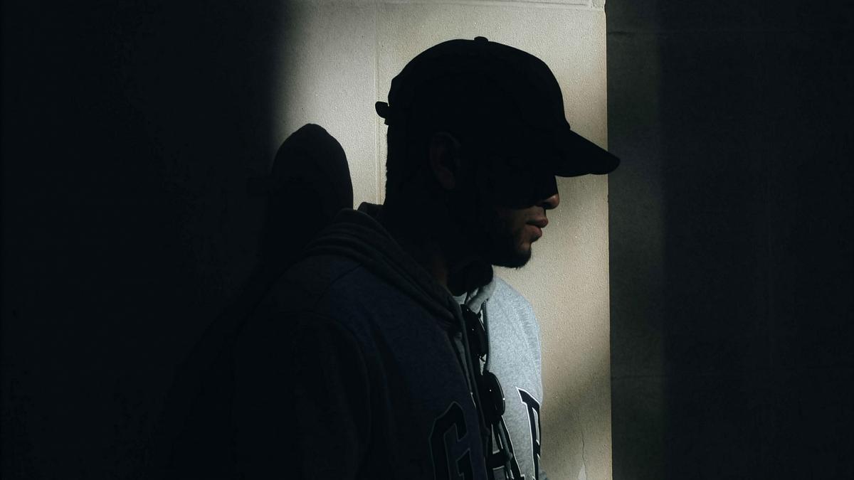 A Black man in a cap and hoodie standing in shadow against a lit wall.