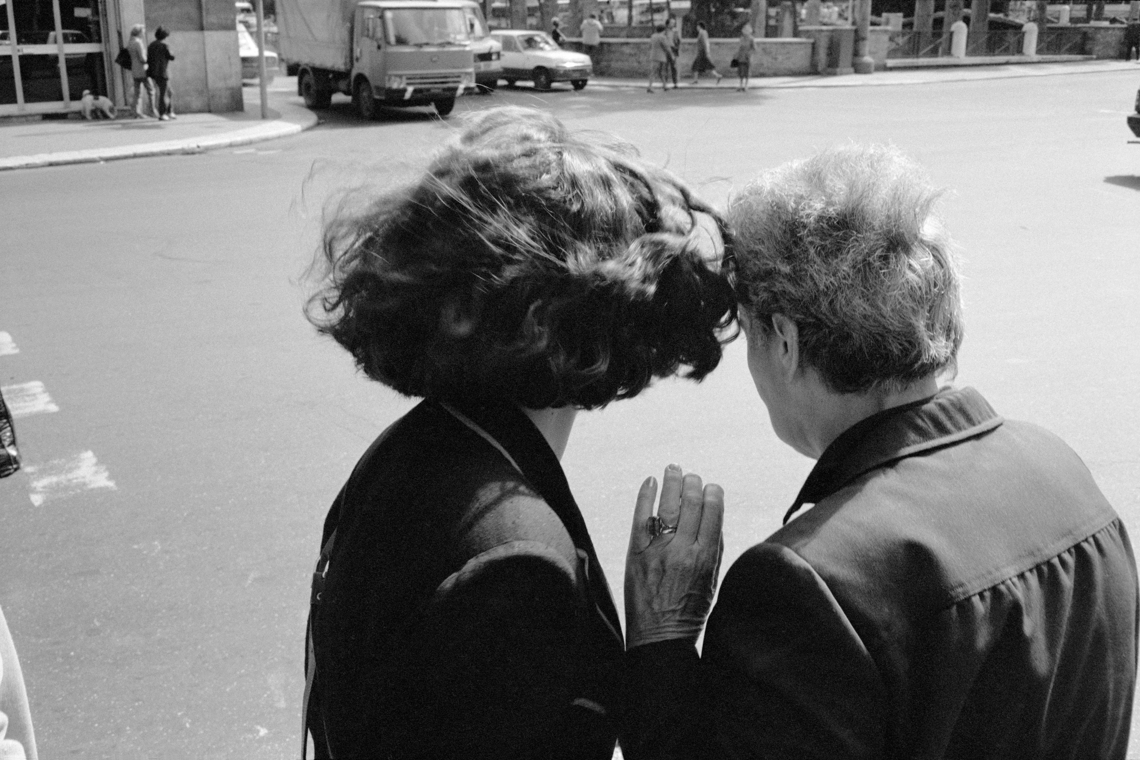 Black and white photo of two people facing away whispering on a street corner with vehicles in the background.