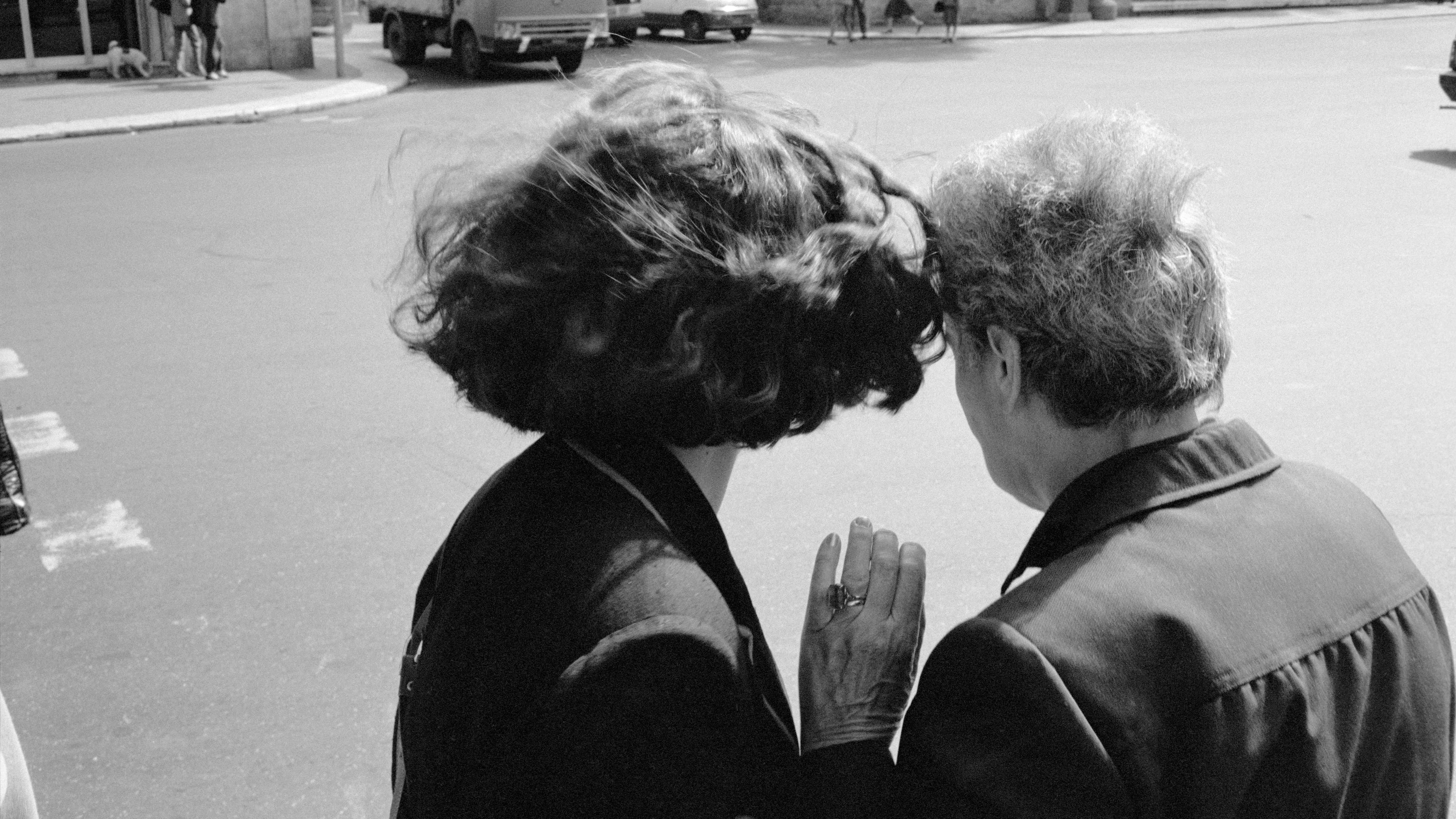 Black and white photo of two people facing away whispering on a street corner with vehicles in the background.