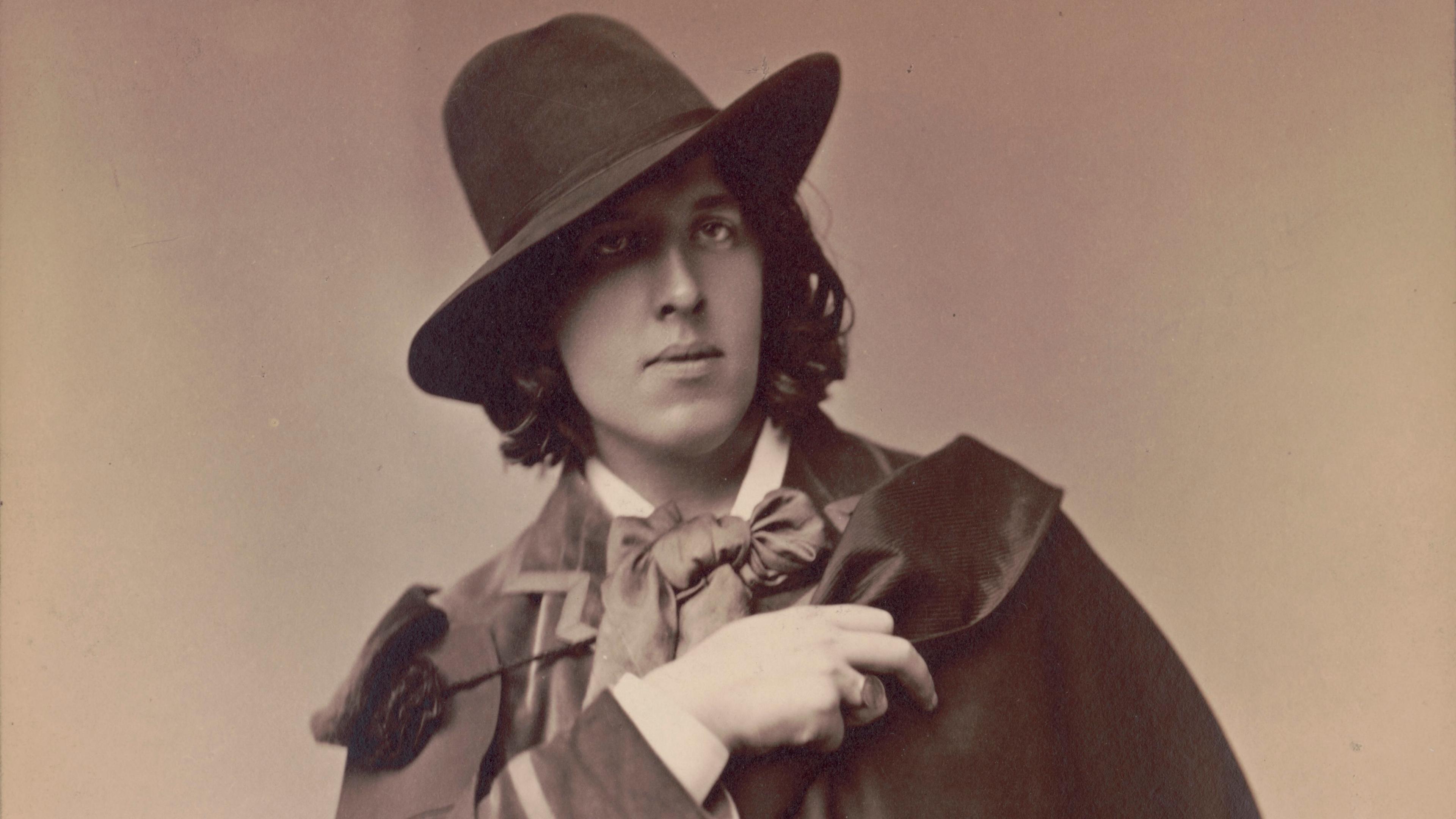 Black and white photo of a person in 19th-century attire with a hat and cape, hand on chest.