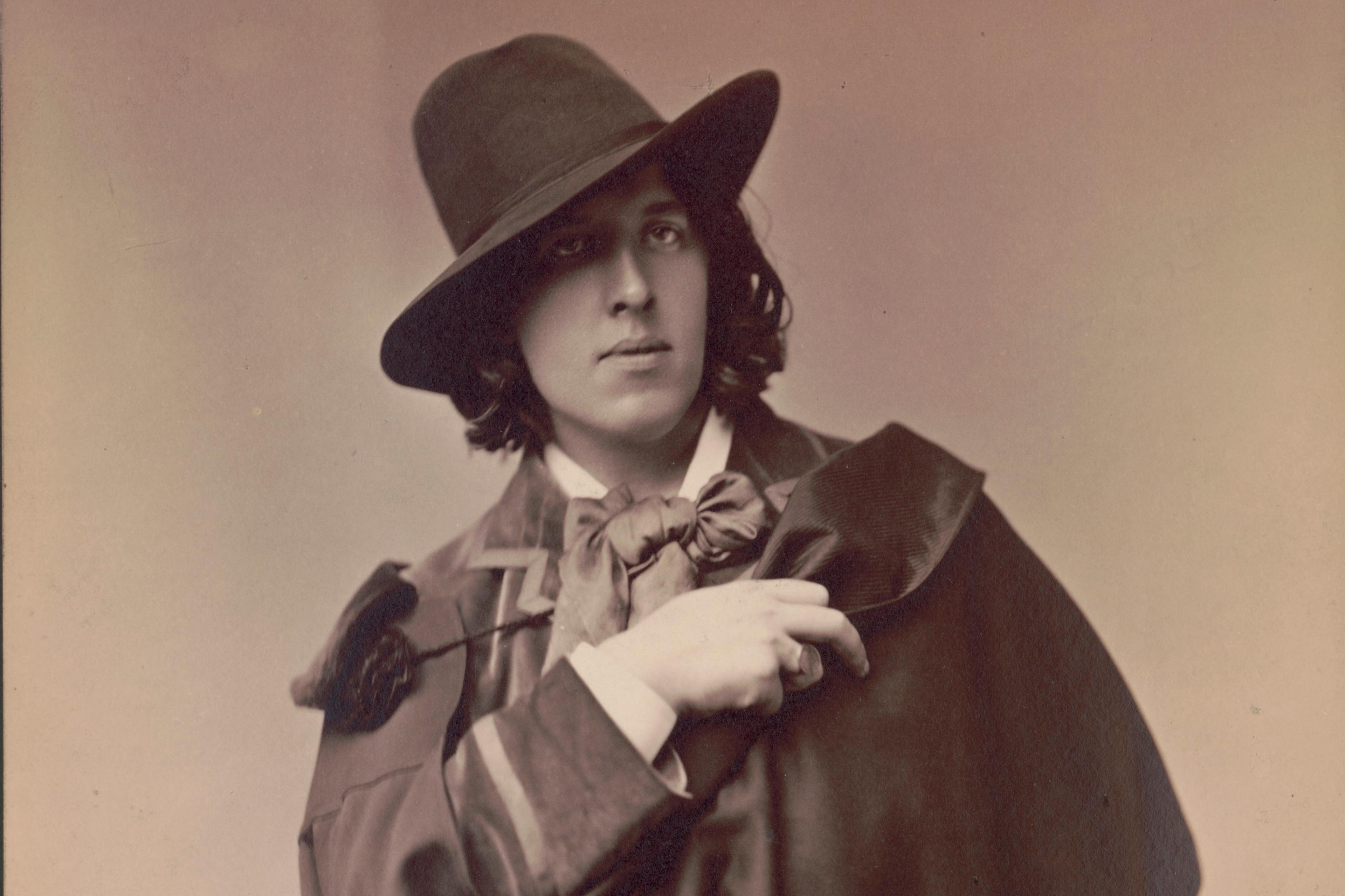 Black and white photo of a person in 19th-century attire with a hat and cape, hand on chest.