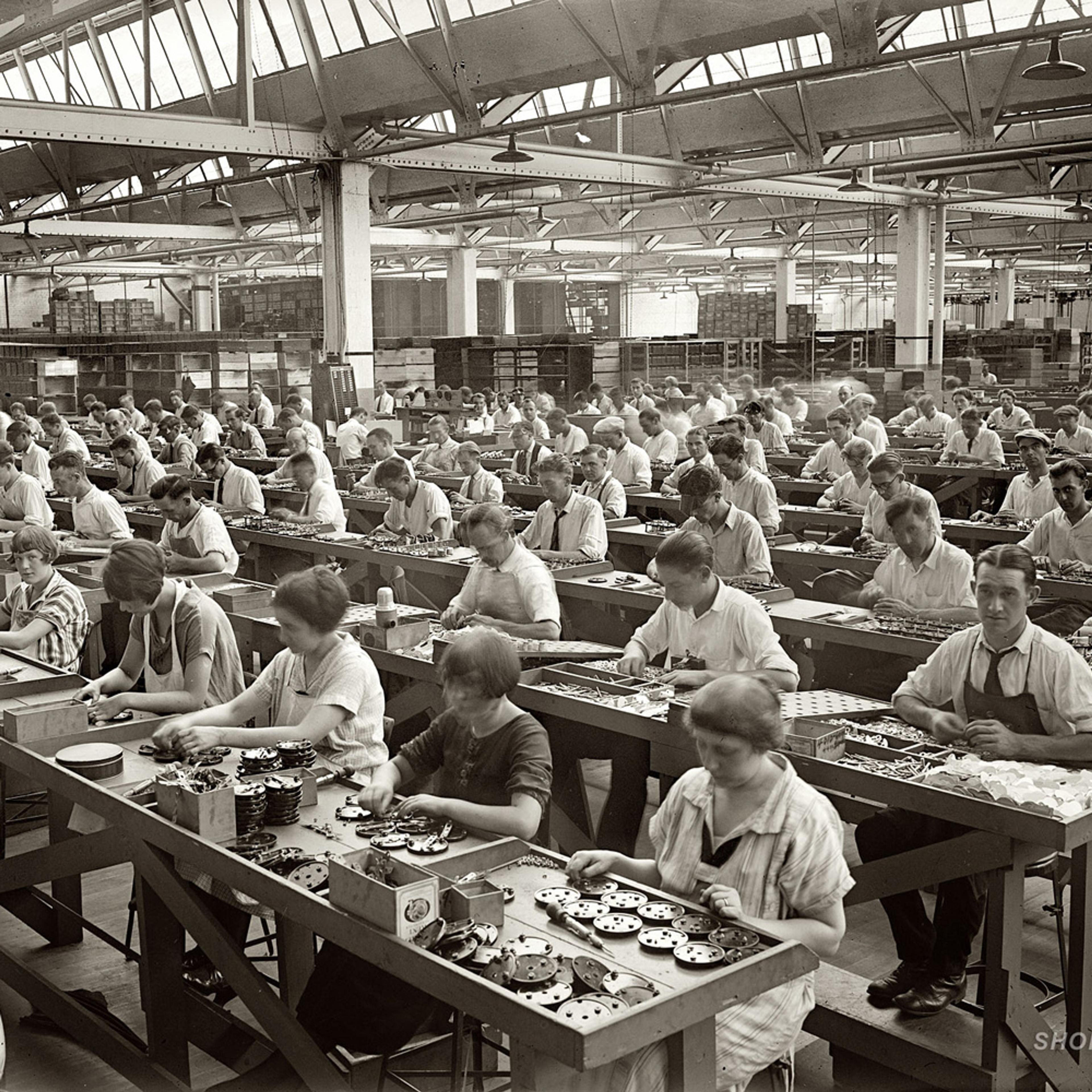 A large factory with numerous workers assembled in rows, working on machinery parts at long wooden tables.