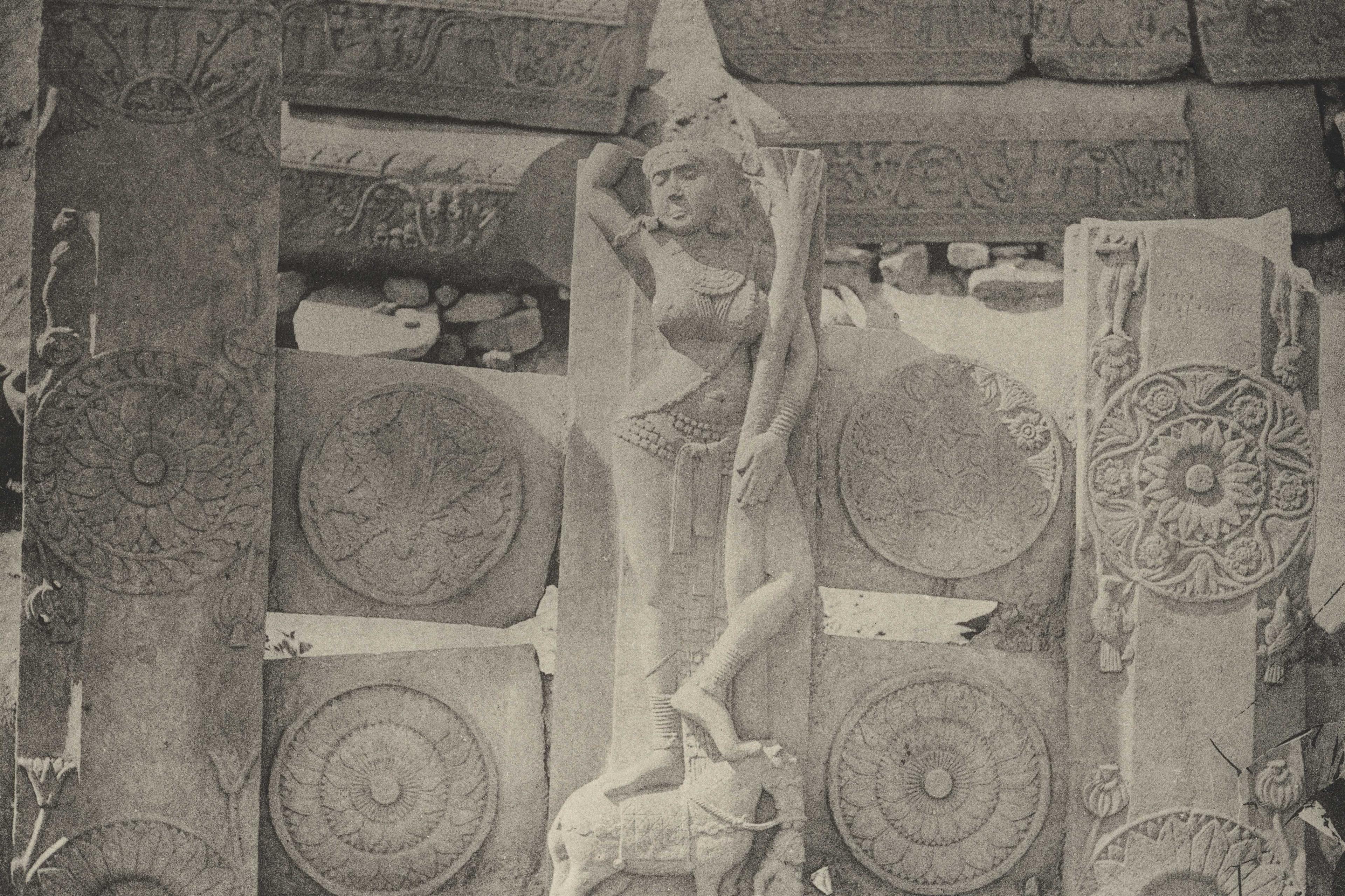 Black and white photo of ancient stone carvings with a central female figure, ornate patterns and a reclining animal.