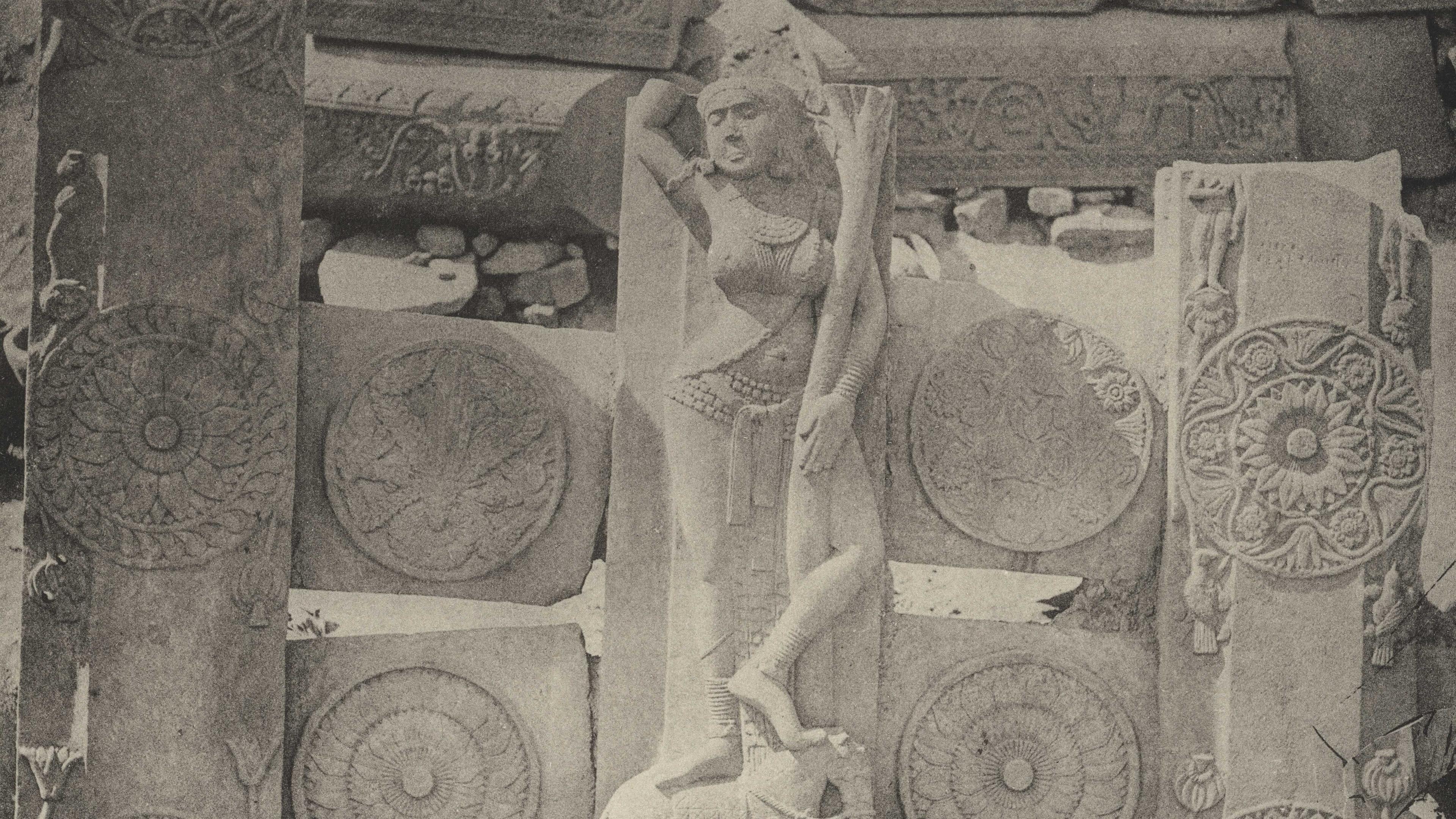 Black and white photo of ancient stone carvings with a central female figure, ornate patterns and a reclining animal.