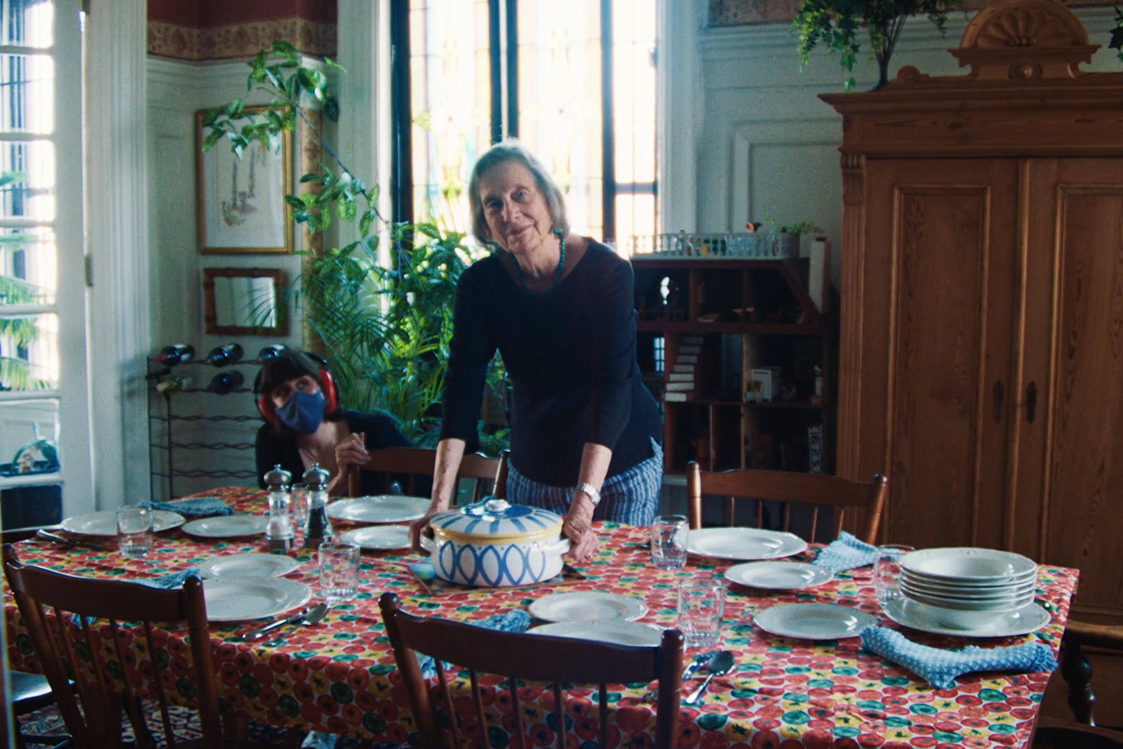 An elder woman setting a dining table with patterned cloth, another person in a mask and headphones in the background, plants, and wooden furniture around.