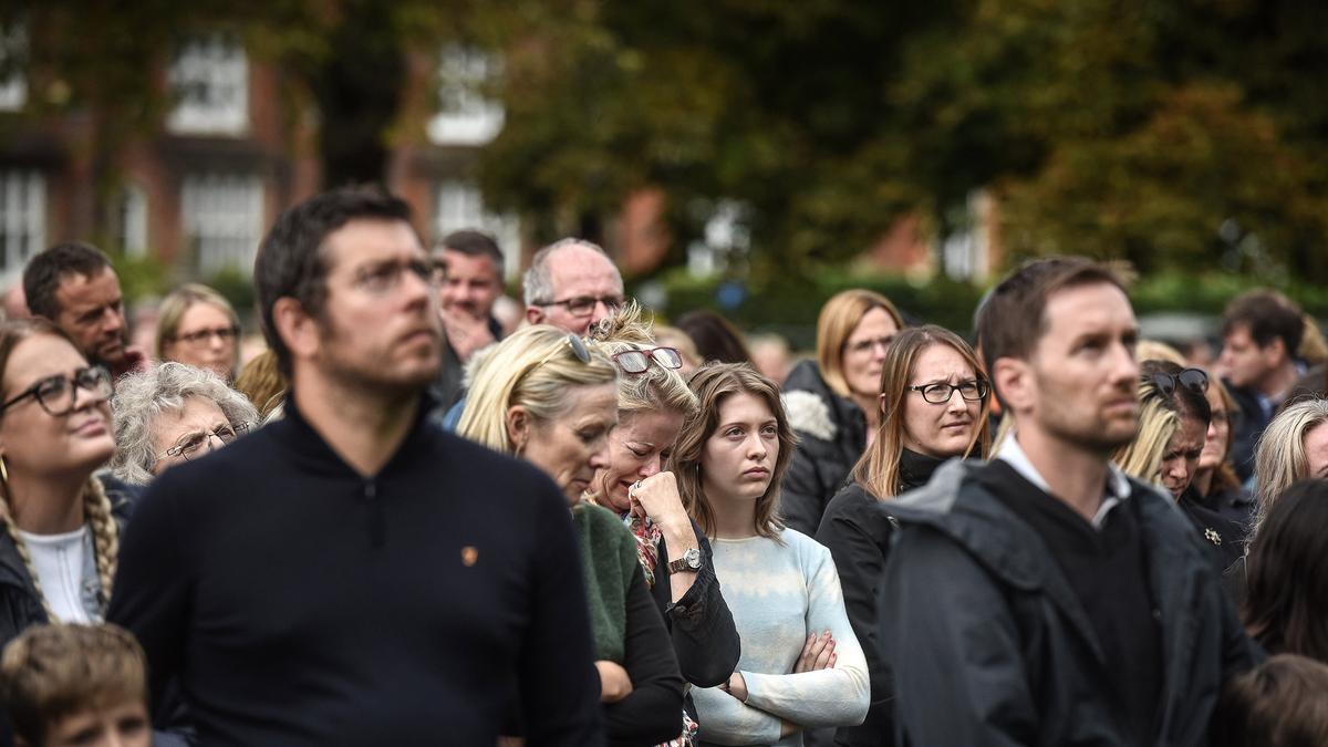 A group of people outdoors looking serious, with one woman wiping her eyes in the centre. Trees and buildings in background.