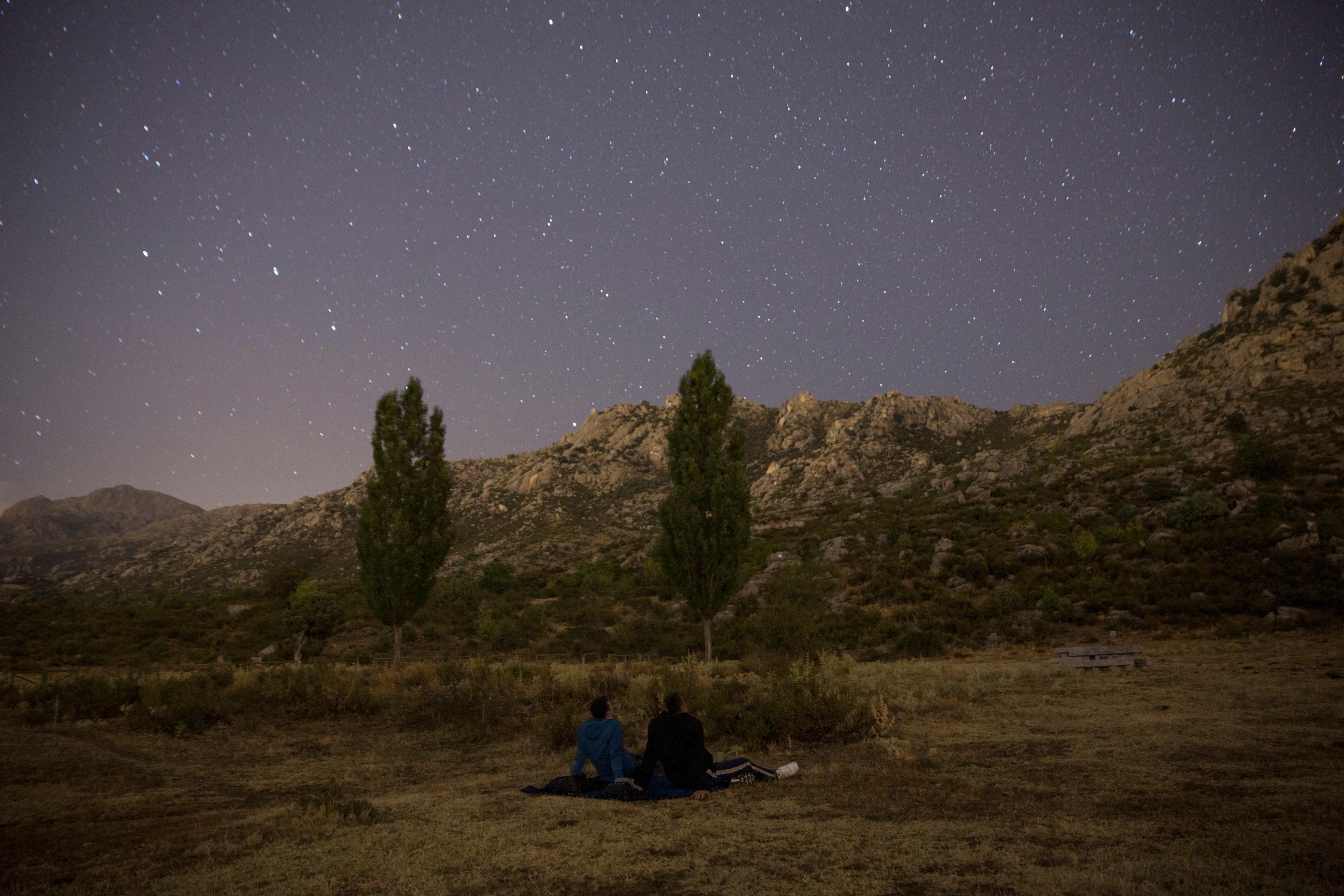 Photo of two people stargazing in a field under a starry sky with mountains and trees in the background.