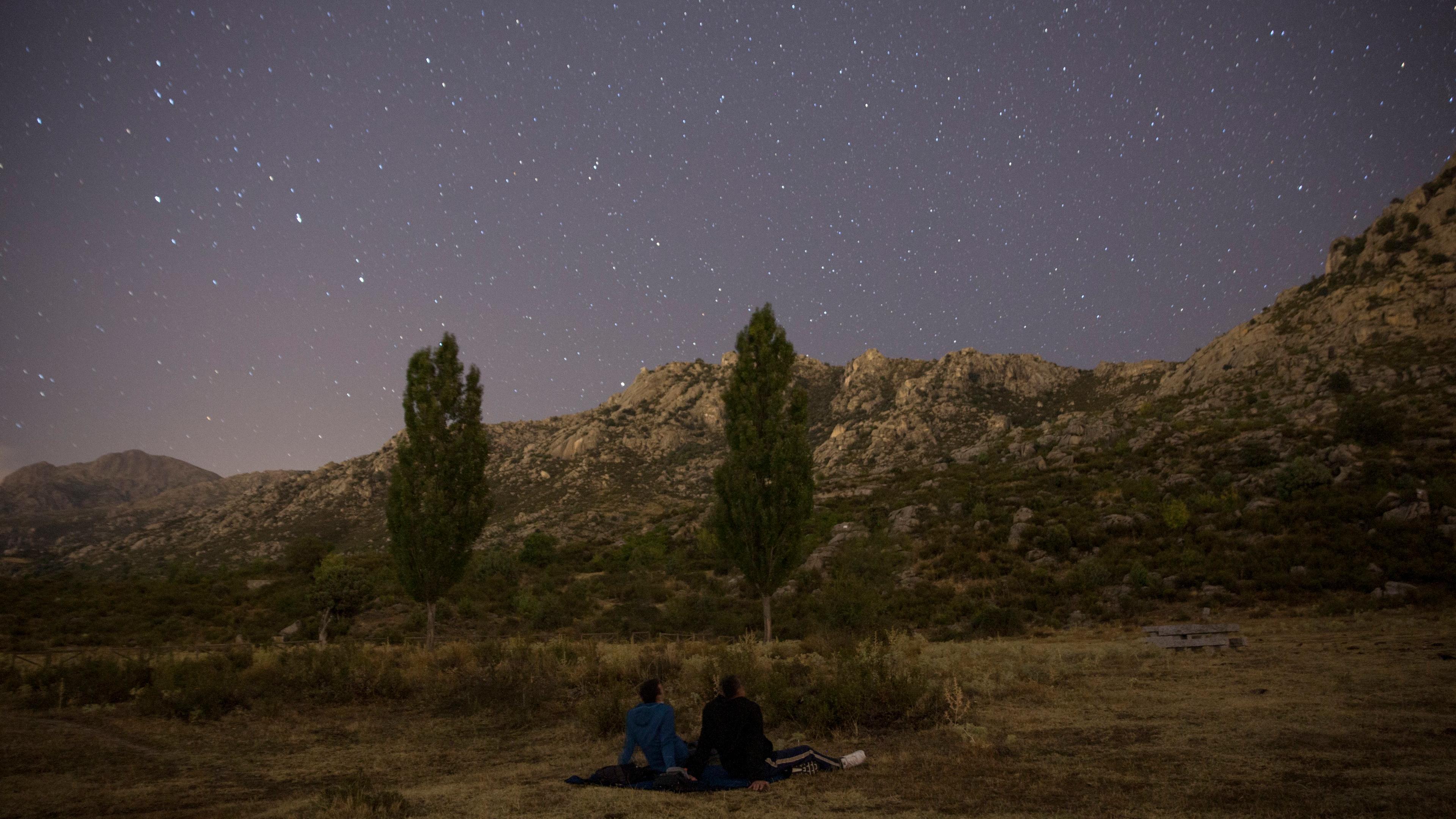 Photo of two people stargazing in a field under a starry sky with mountains and trees in the background.