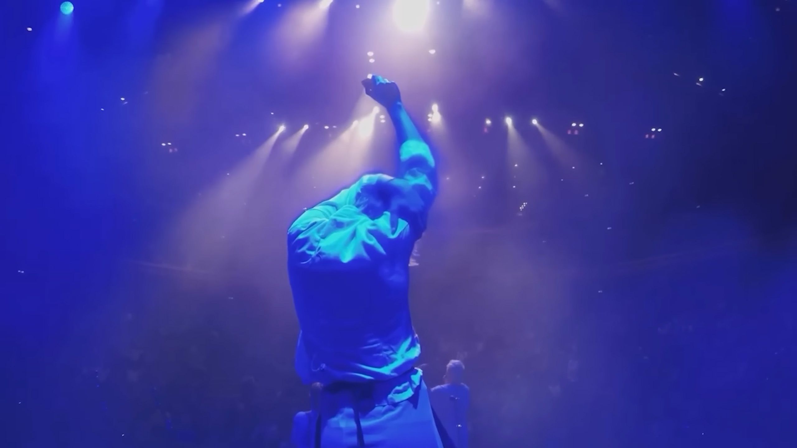 Photo of a person on stage under blue lights with an arm raised, facing an unseen audience in a smoky atmosphere.