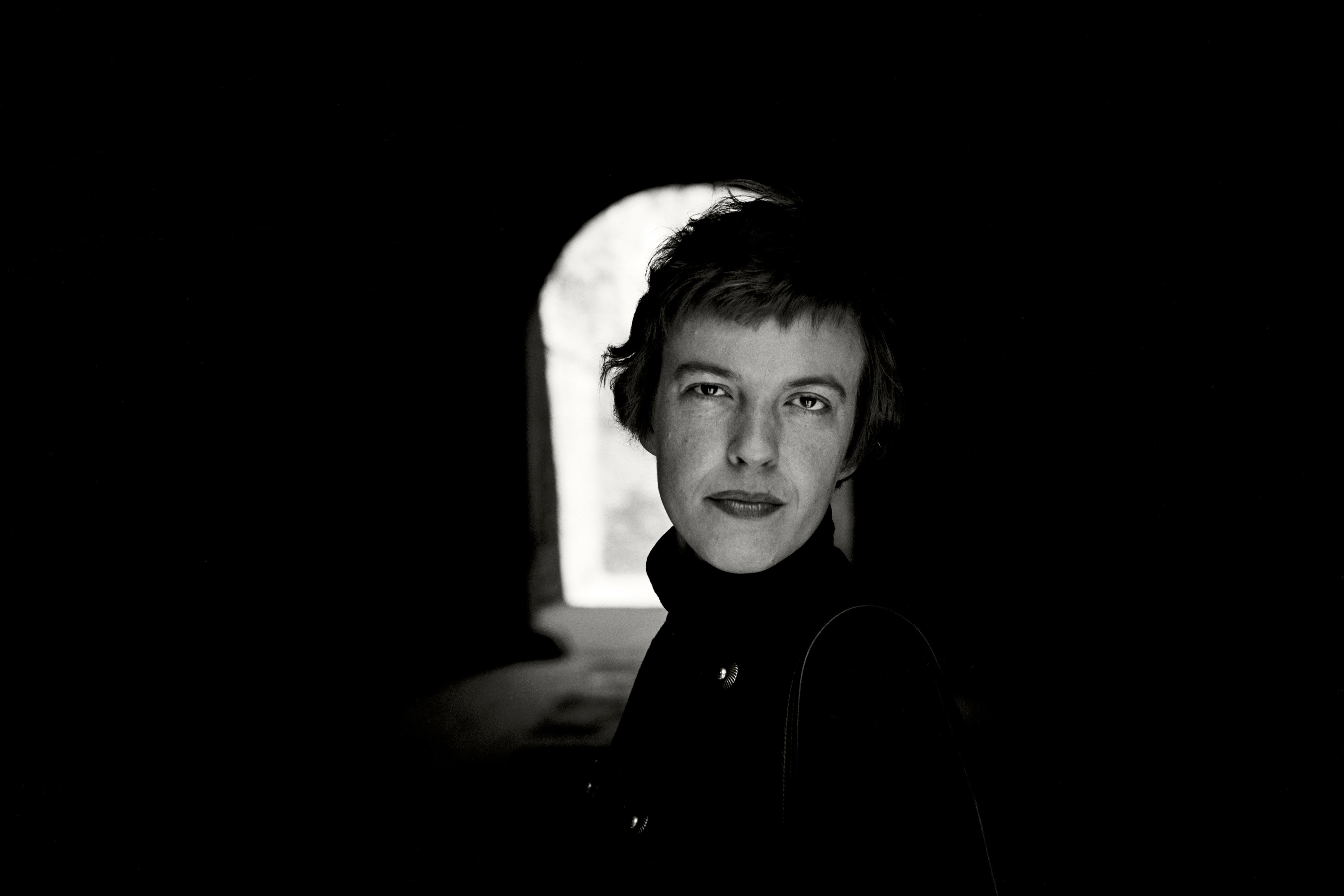 Black and white photo of a person in a dark tunnel with light in the background, wearing a coat, looking at the camera.